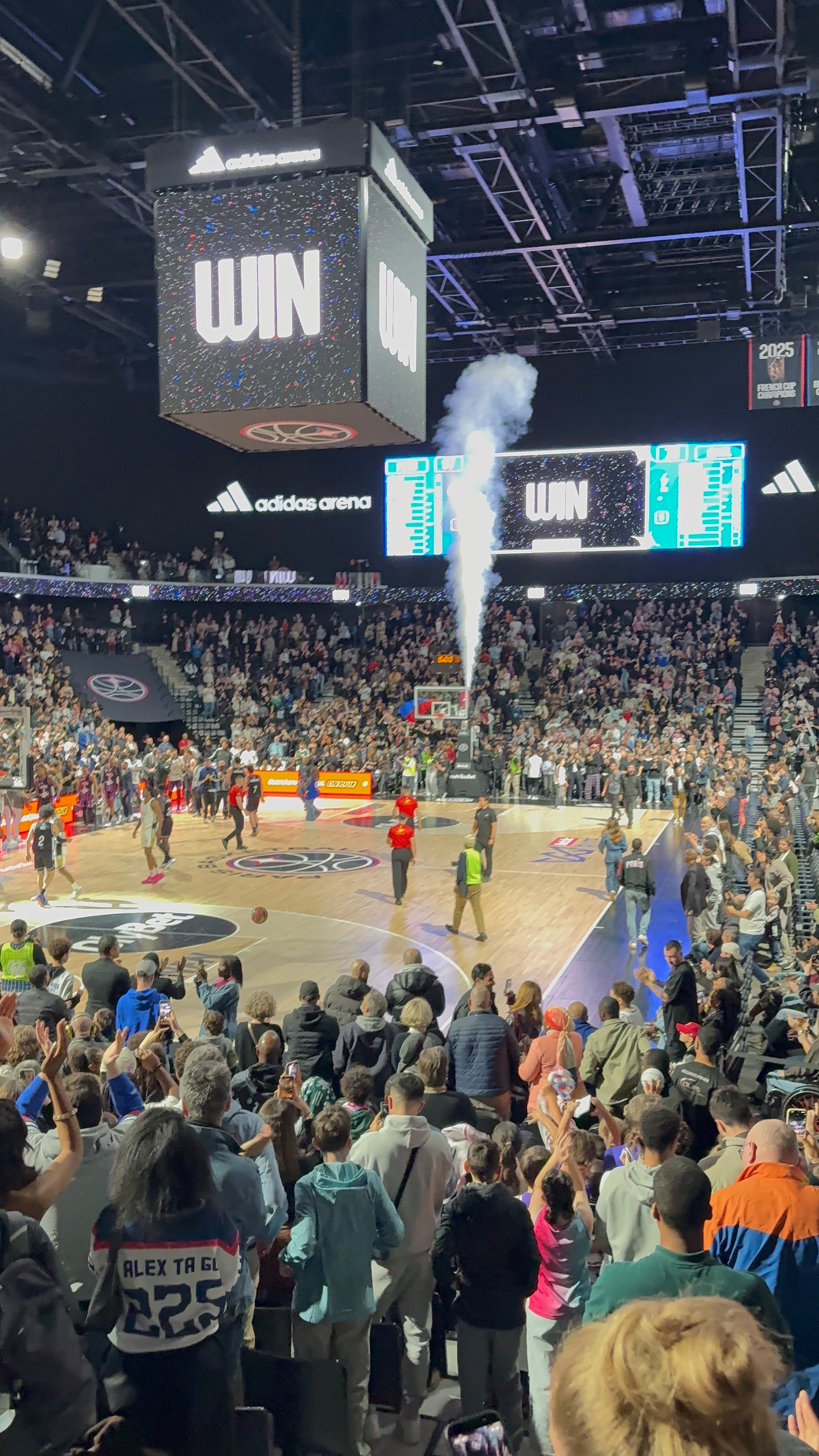 A packed basketball arena celebrates a team's victory with smoke effects and a WIN displayed on the screen.