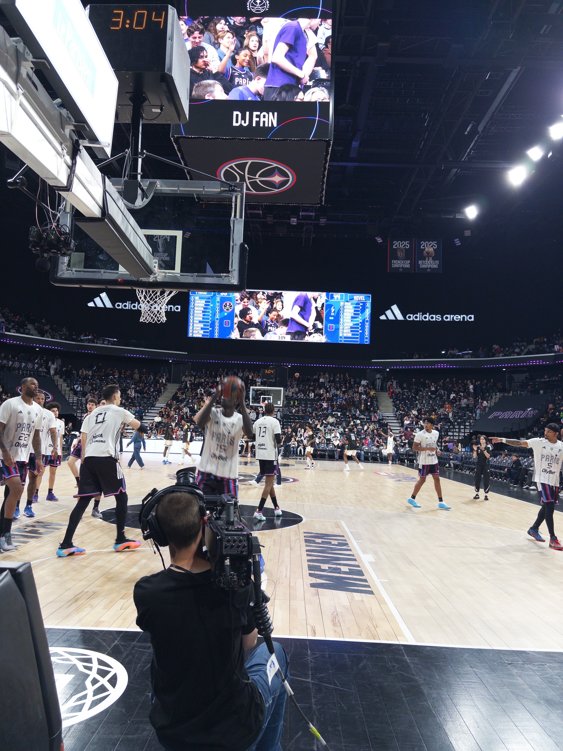 Basketball players are warming up on a court while a cameraman films the action, surrounded by a large audience in an indoor arena.