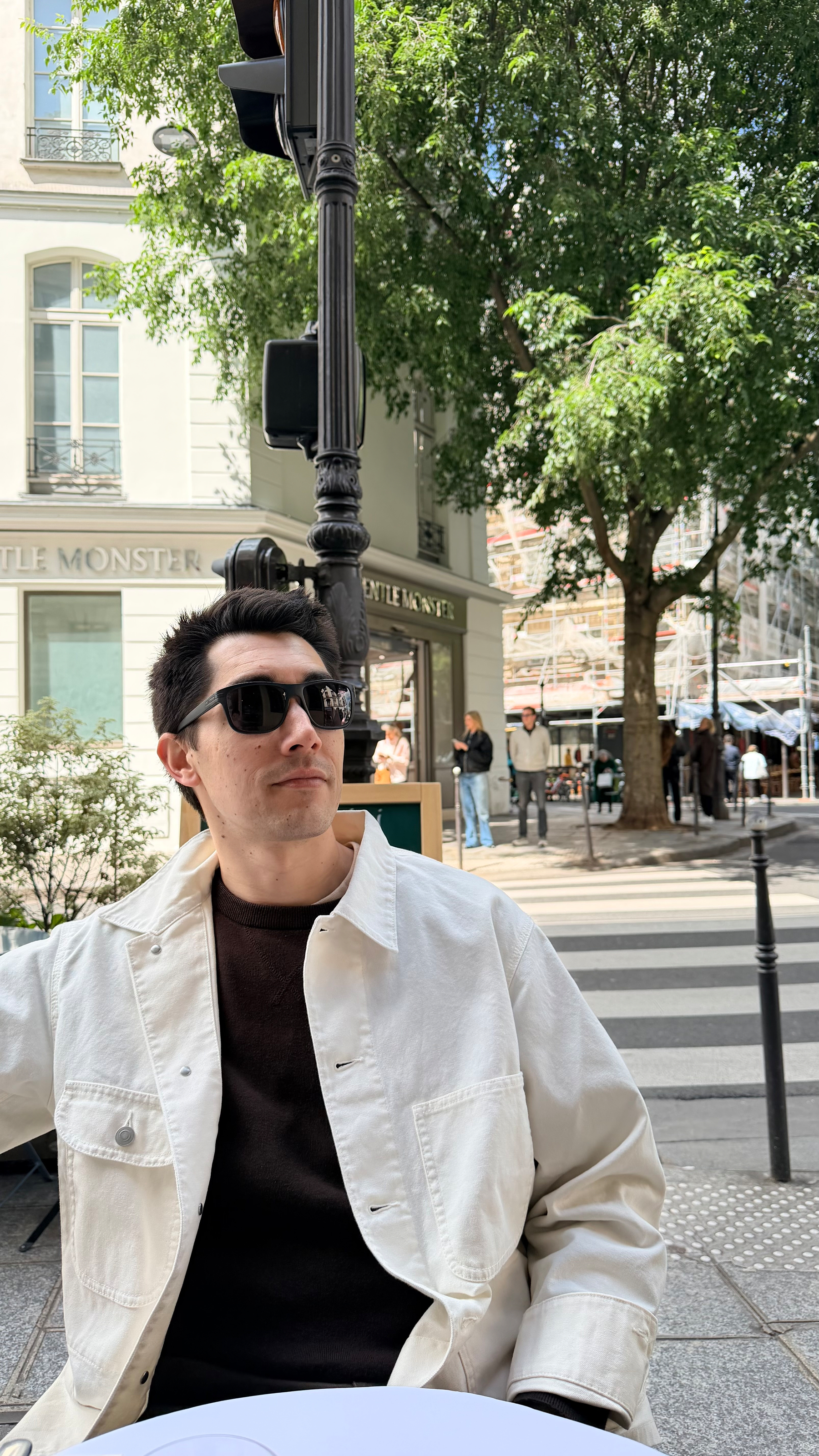 Auto-generated description: A person wearing sunglasses and a white jacket is sitting at an outdoor café on a sunny street with trees and buildings in the background.