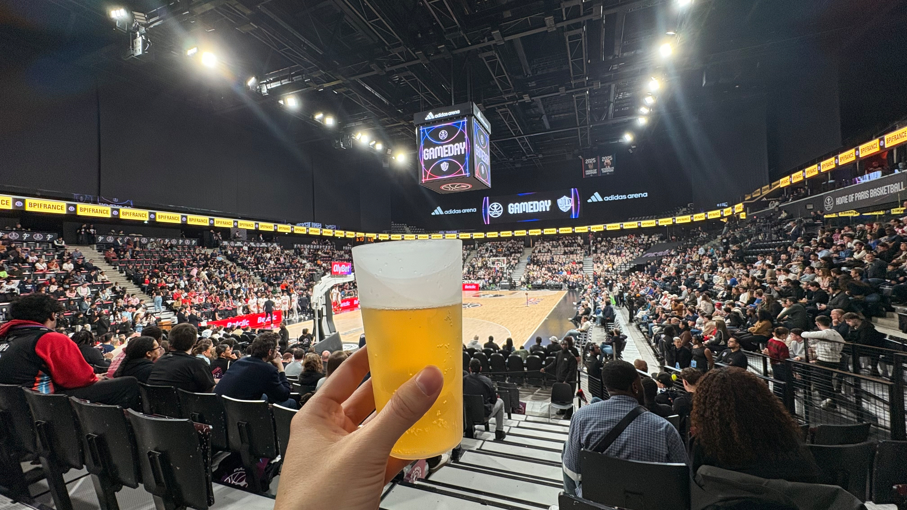 A person is holding a cup of beer while seated in an arena watching a basketball game.