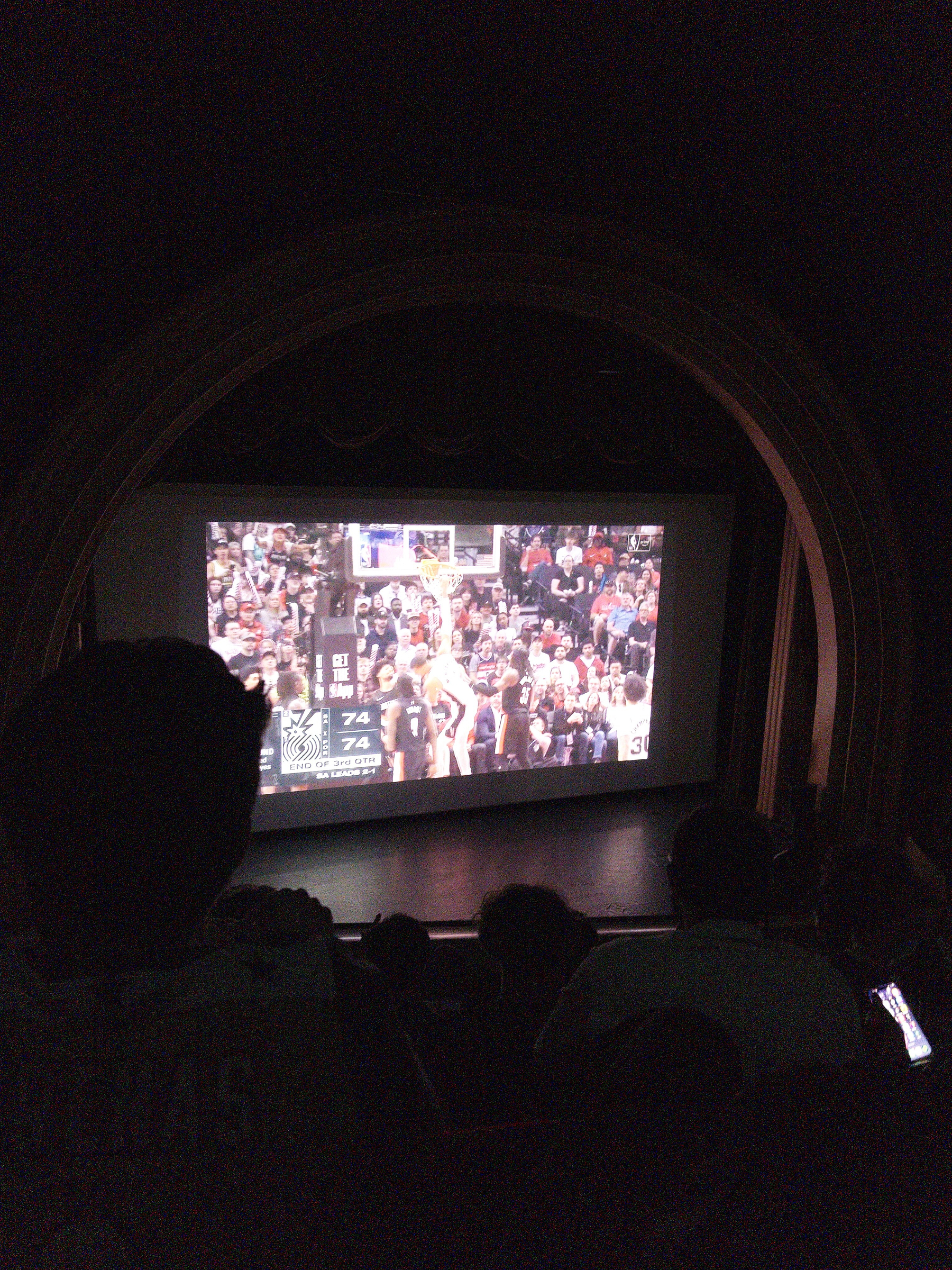 A large indoor screen displays a basketball game to an audience seated in a theater.
