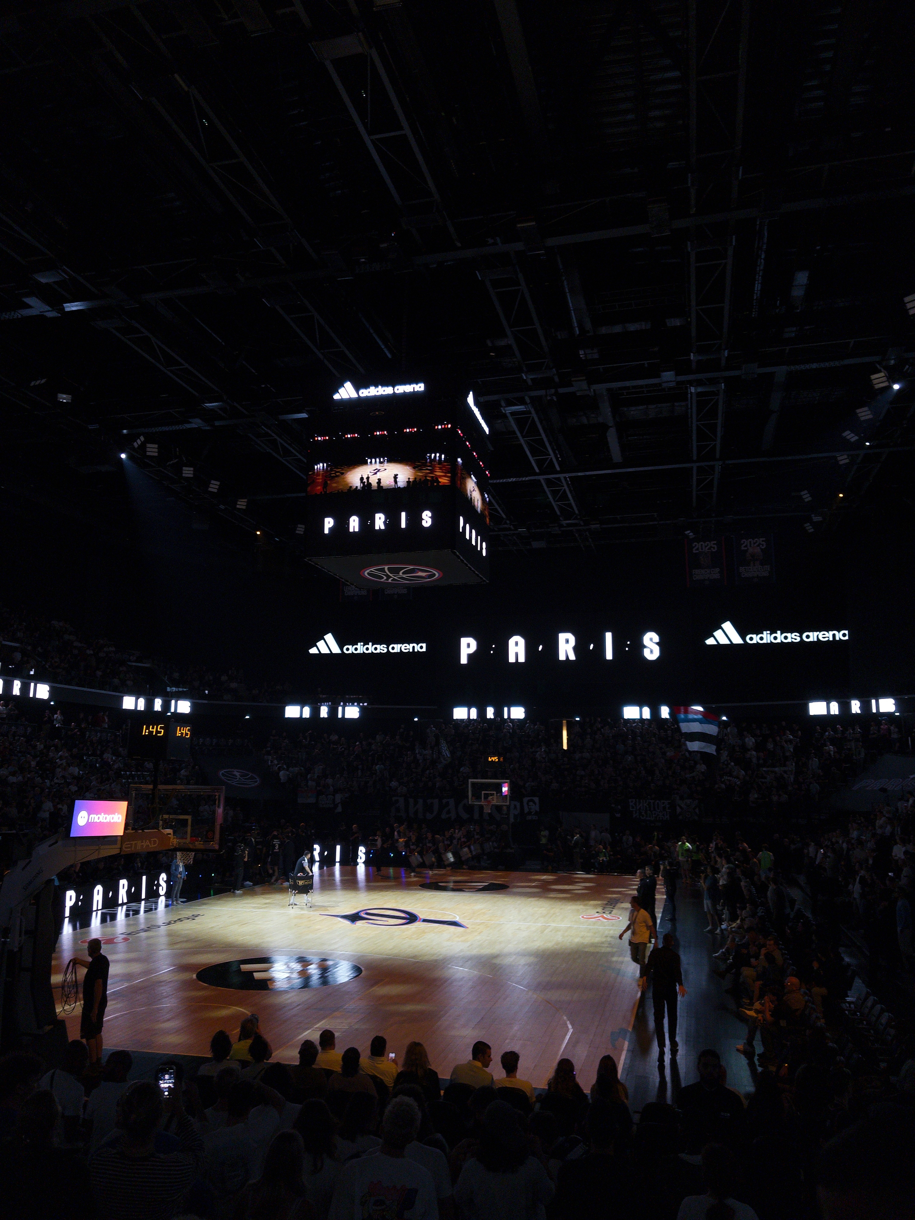 A basketball court inside an arena with PARIS and adidas arena displayed prominently on the scoreboard and digital screens.
