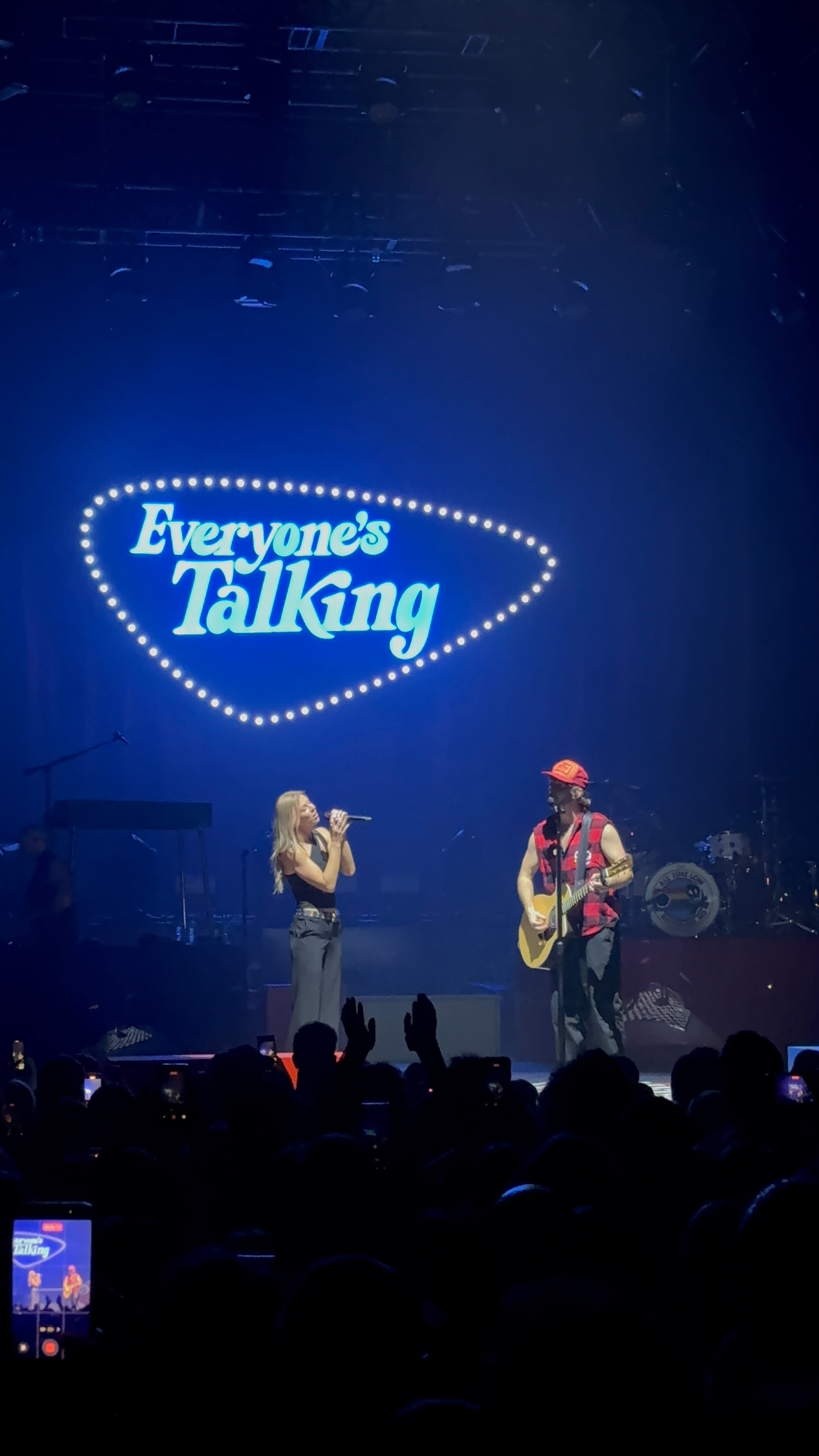 Two performers are on stage under a sign that says Everyone's Talking, with the audience capturing the moment on their phones.