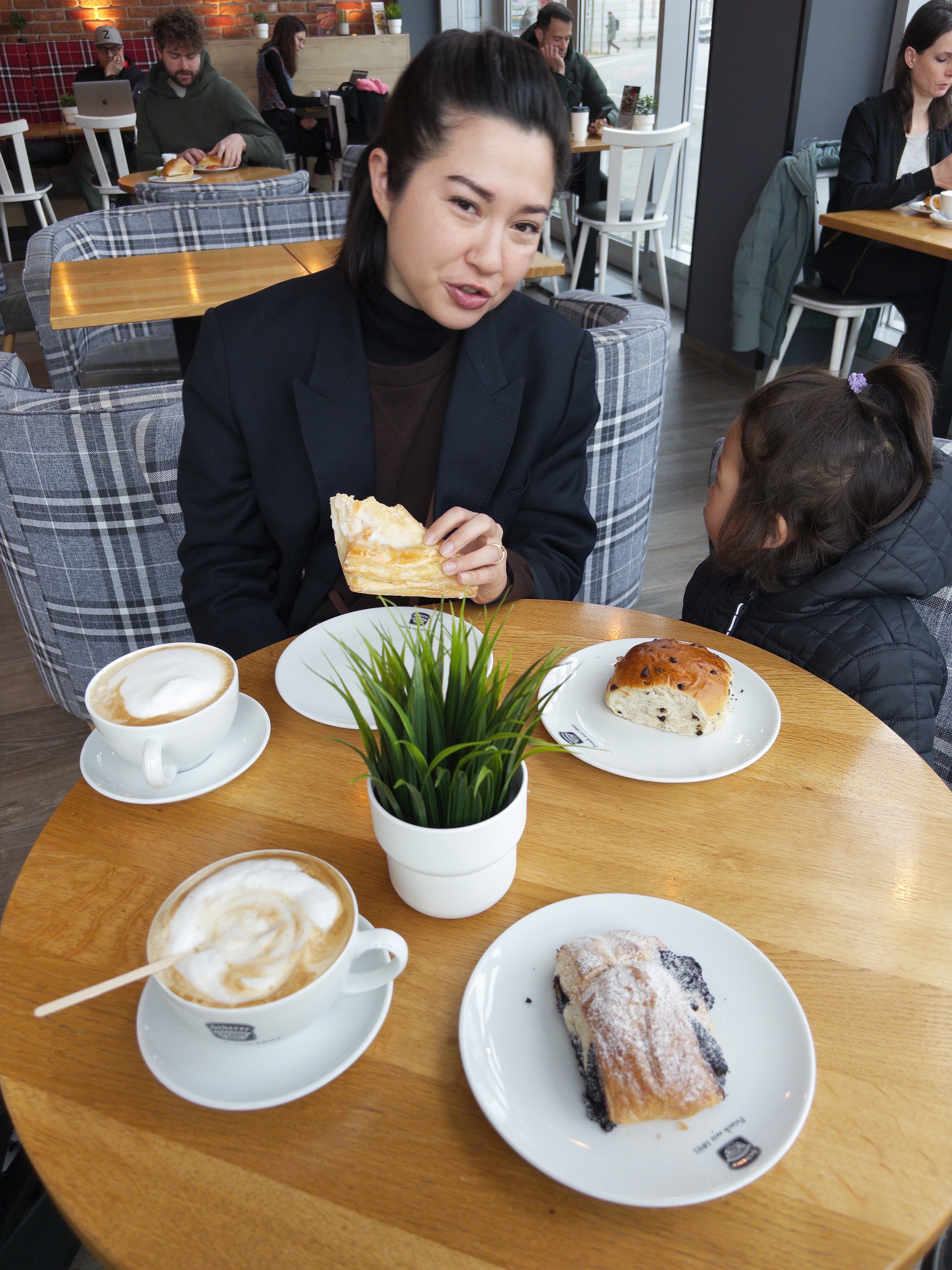 Auto-generated description: A woman and a child are sitting at a café table with pastries, two cups of coffee, and a small plant.