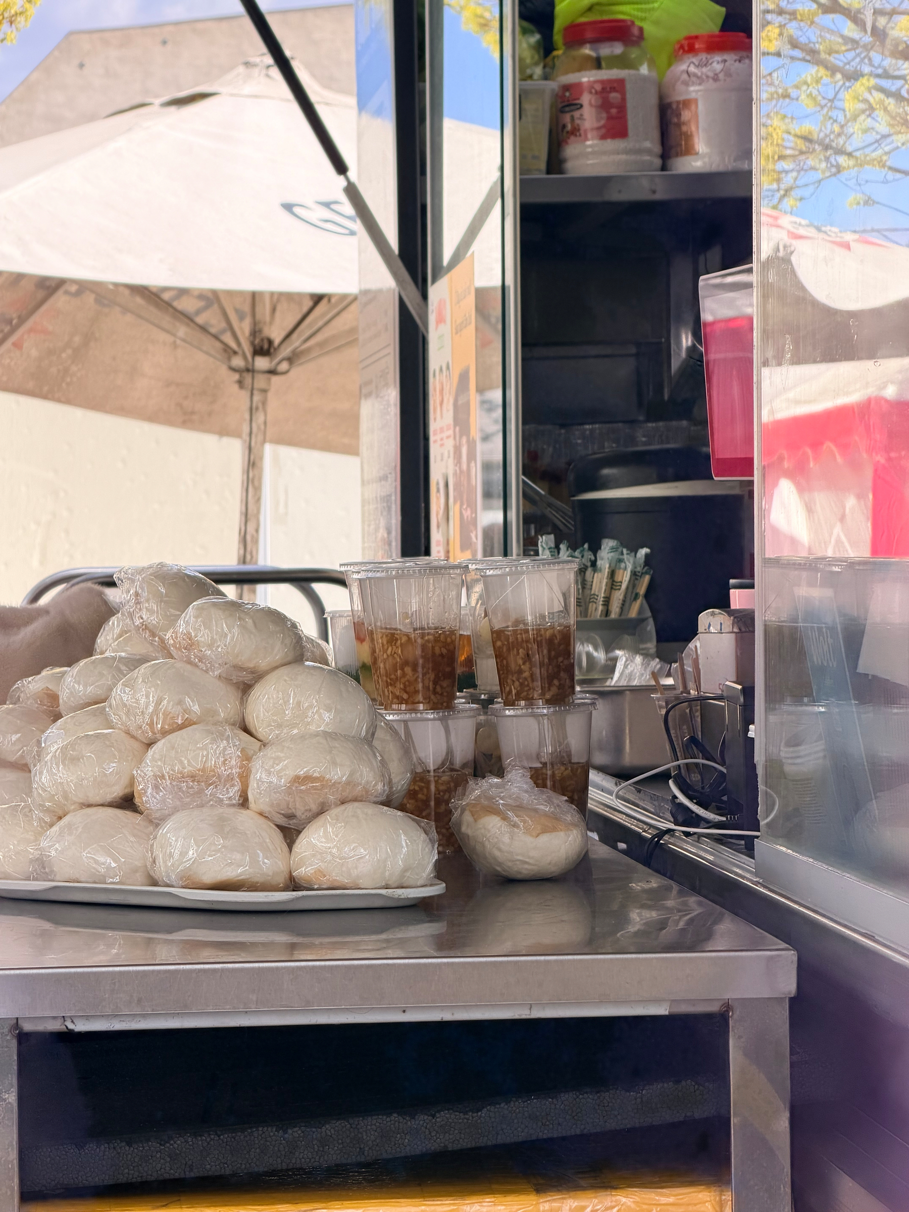 Auto-generated description: A street food stall displays a tray of wrapped buns alongside cups filled with a brown liquid, under an outdoor umbrella.