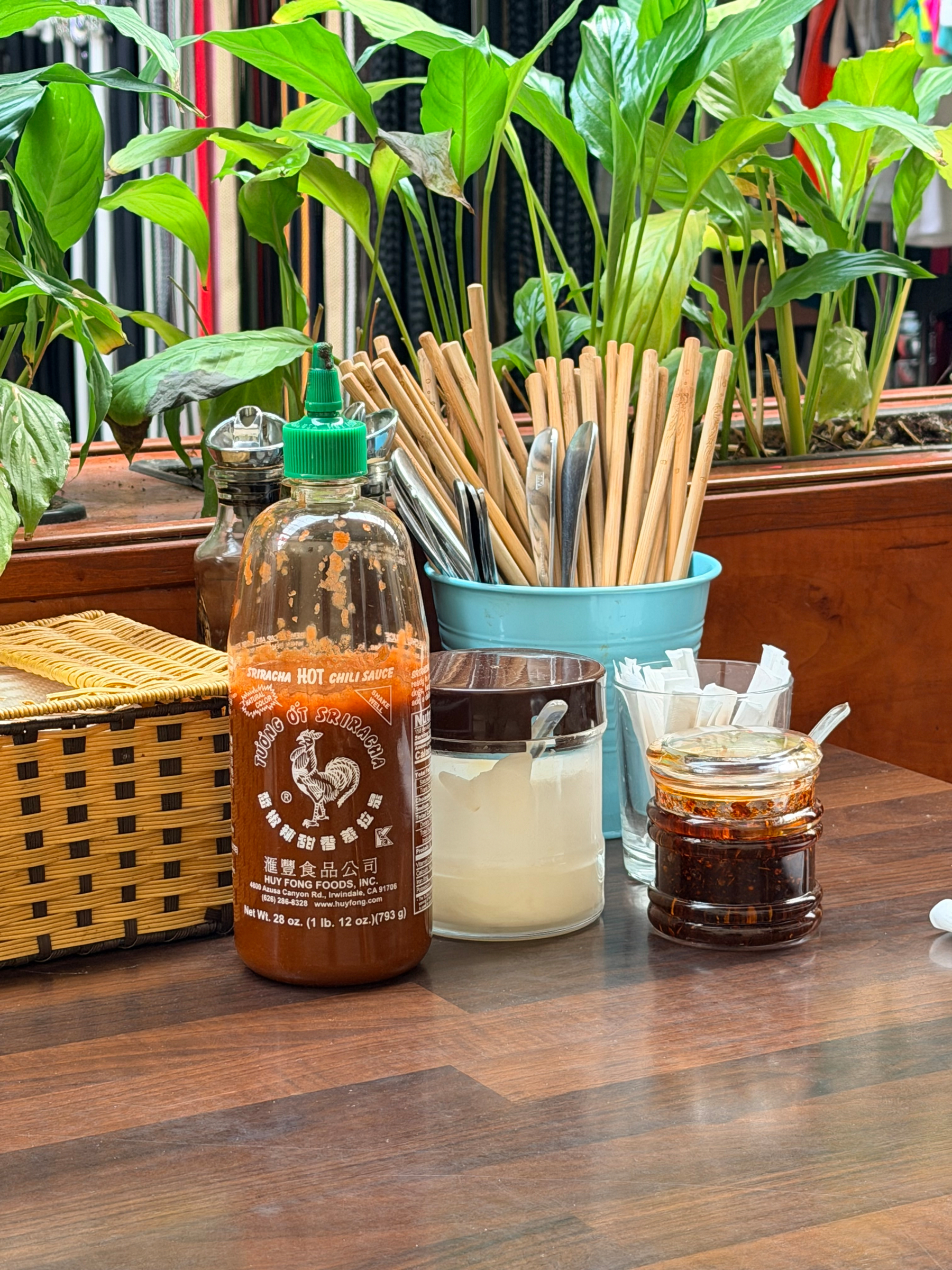 Auto-generated description: A table is set with bottles of condiments, chopsticks in a container, and a basket, surrounded by leafy plants.