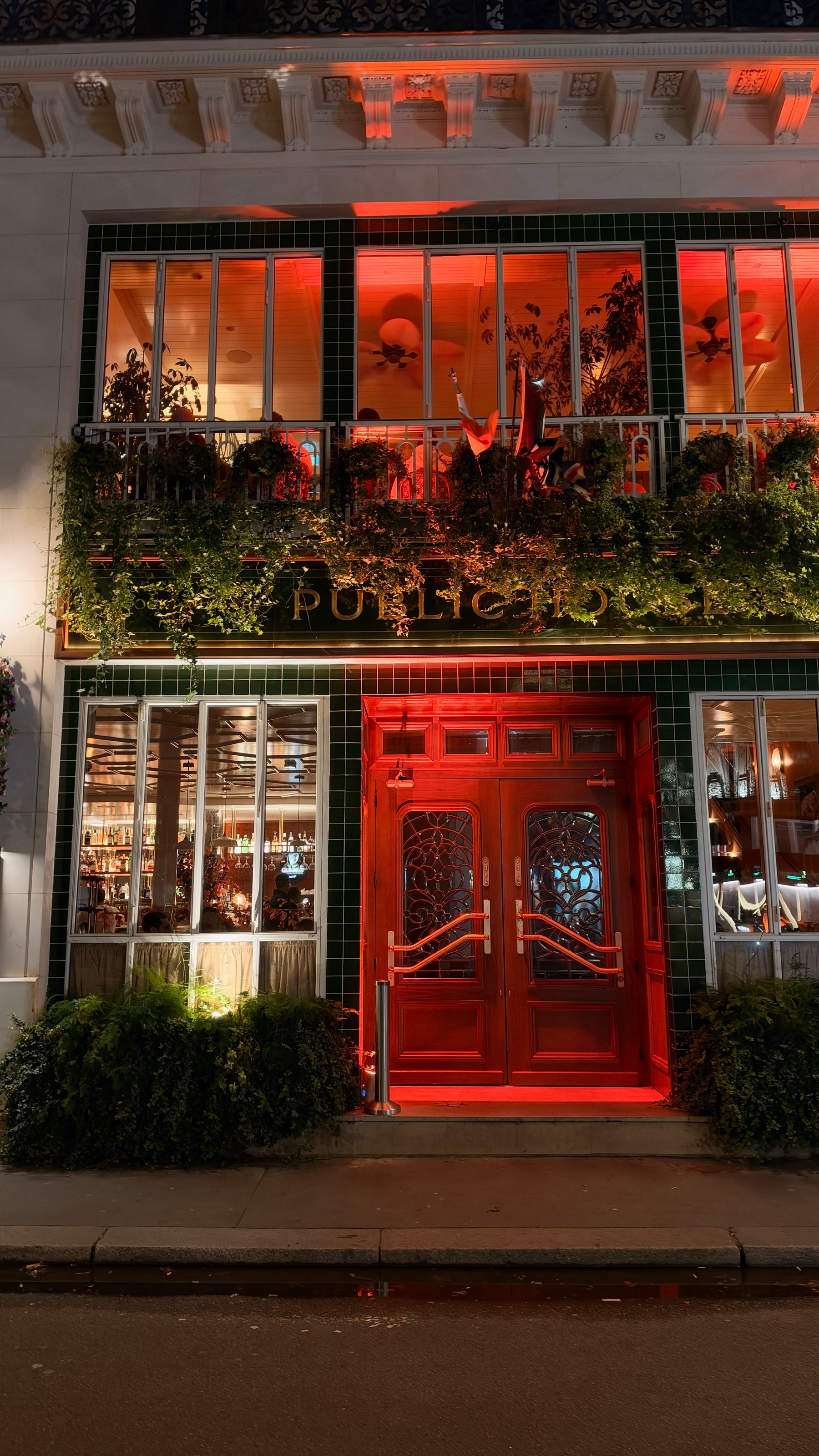 A building with a vibrant red double door entrance and illuminated upper windows framed by green tiles and lush greenery is shown at night.
