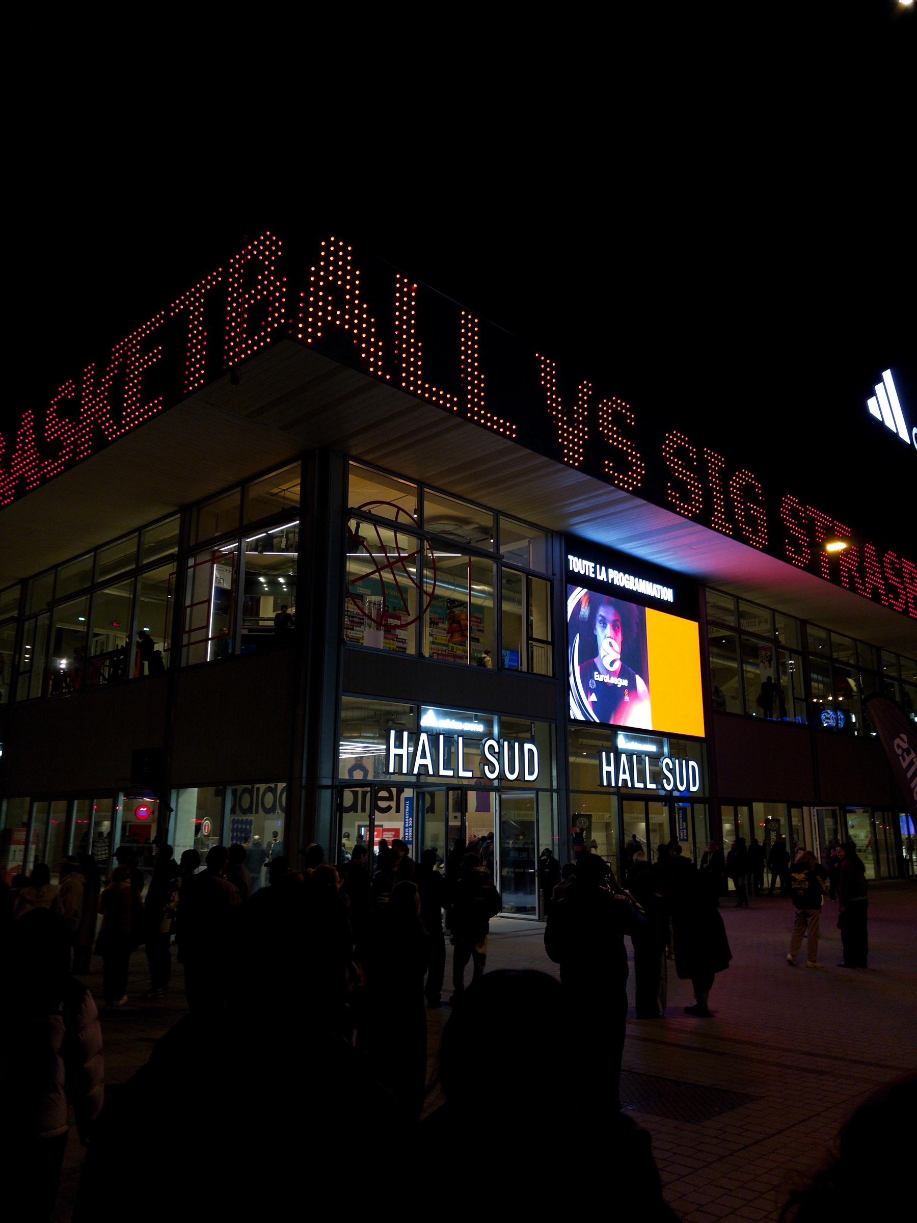 A group of people is gathered outside a brightly lit sports arena with a large sign displaying Paris Basketball vs SIG Strasbourg.