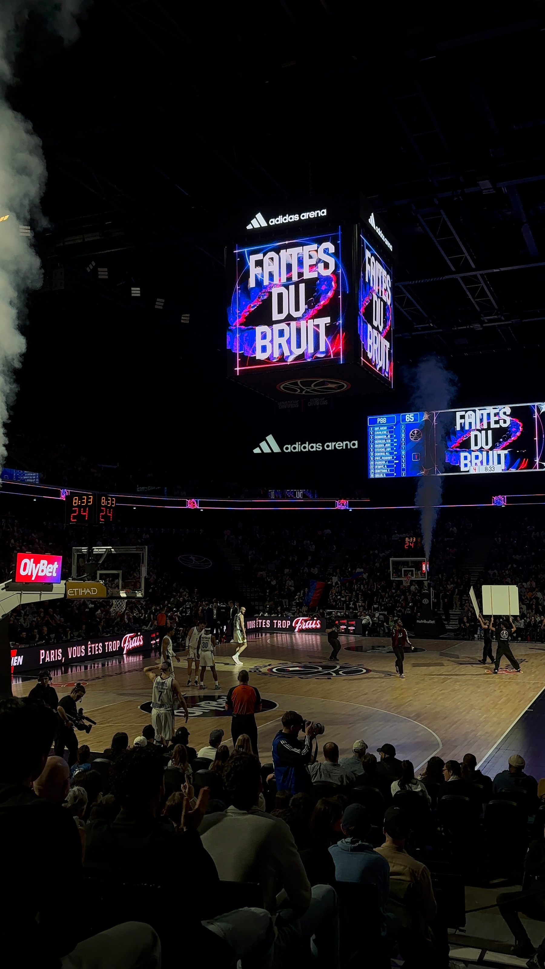 A basketball game is taking place in a large arena with a scoreboard displaying vivid graphics and the words FAITES DU BRUIT.