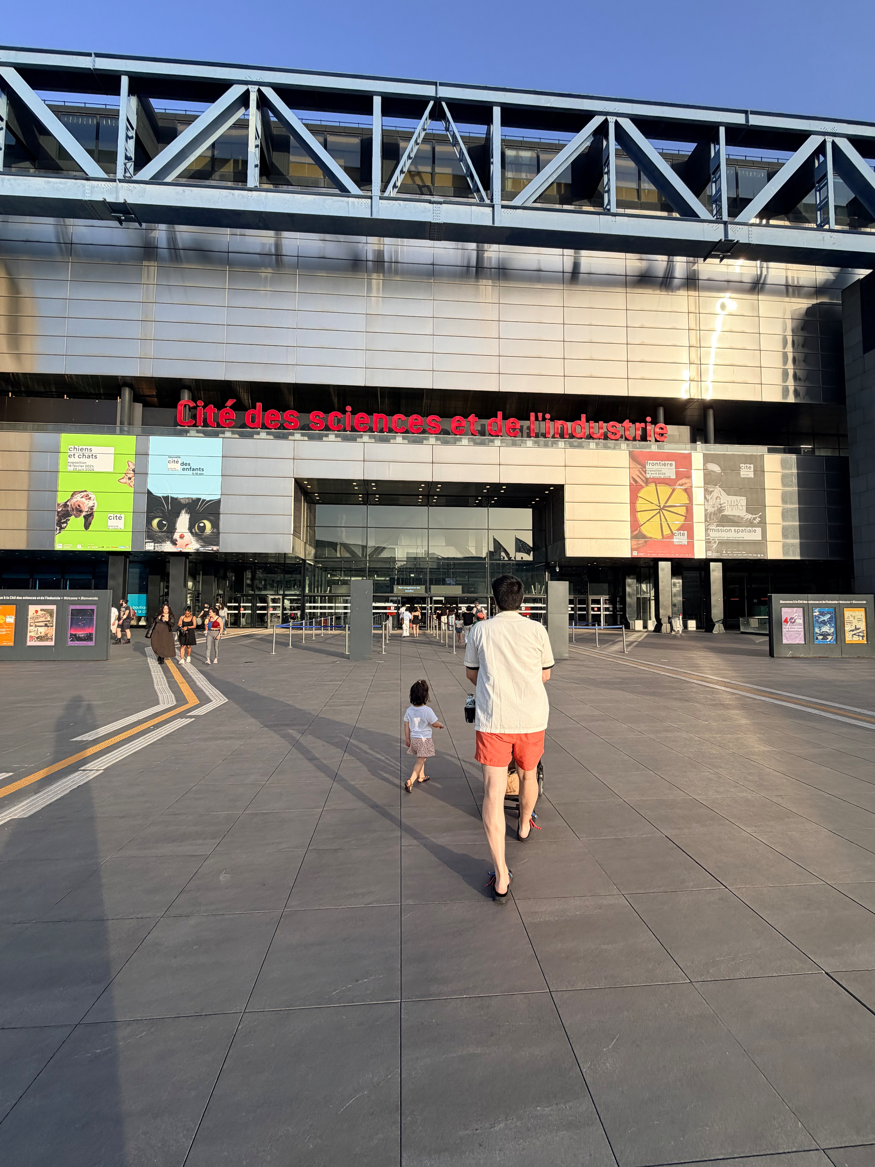A person and a child walk towards the entrance of Cité des Sciences et de l'Industrie in Paris, with colorful posters displayed on the building.