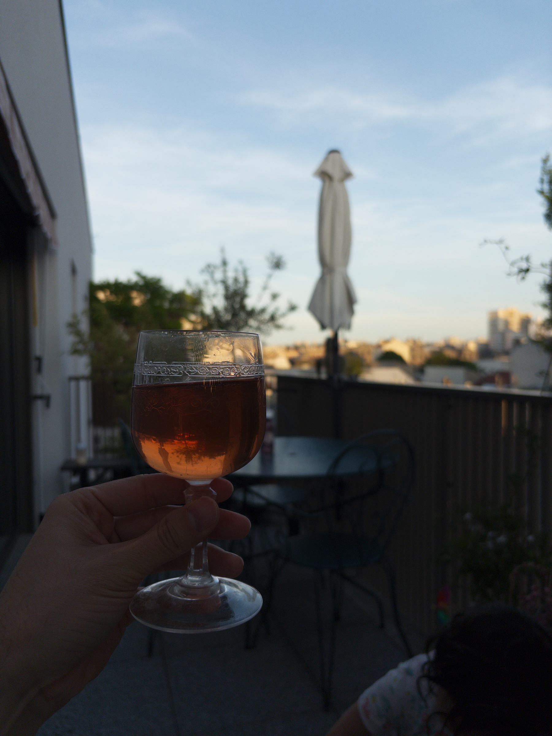 A hand holds a glass of rosé wine on a balcony with outdoor seating, overlooking a view under a clear sky.