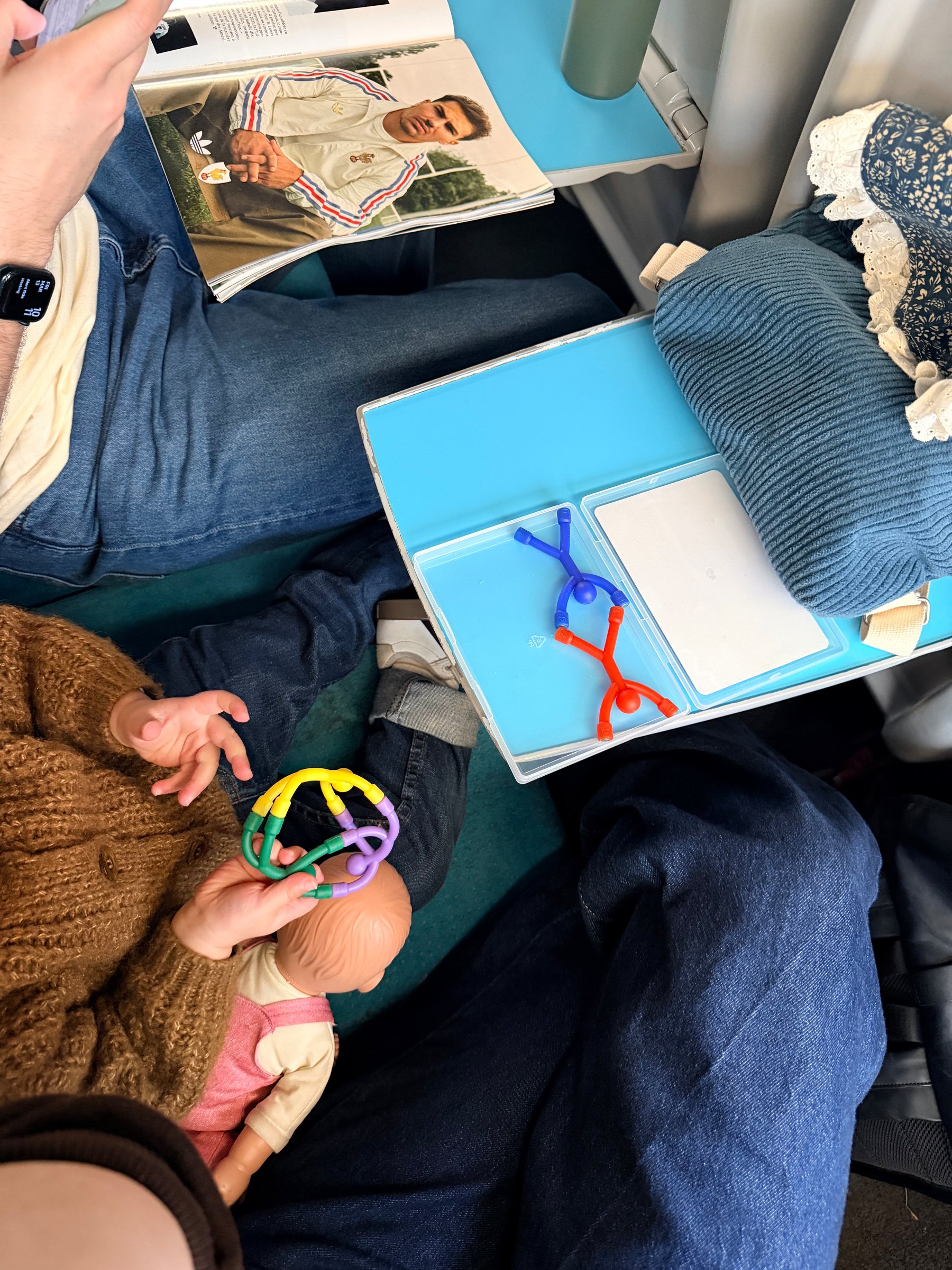 Auto-generated description: A child holding a colorful toy sits next to an adult reading a magazine, with another adult nearby and items on a small table.