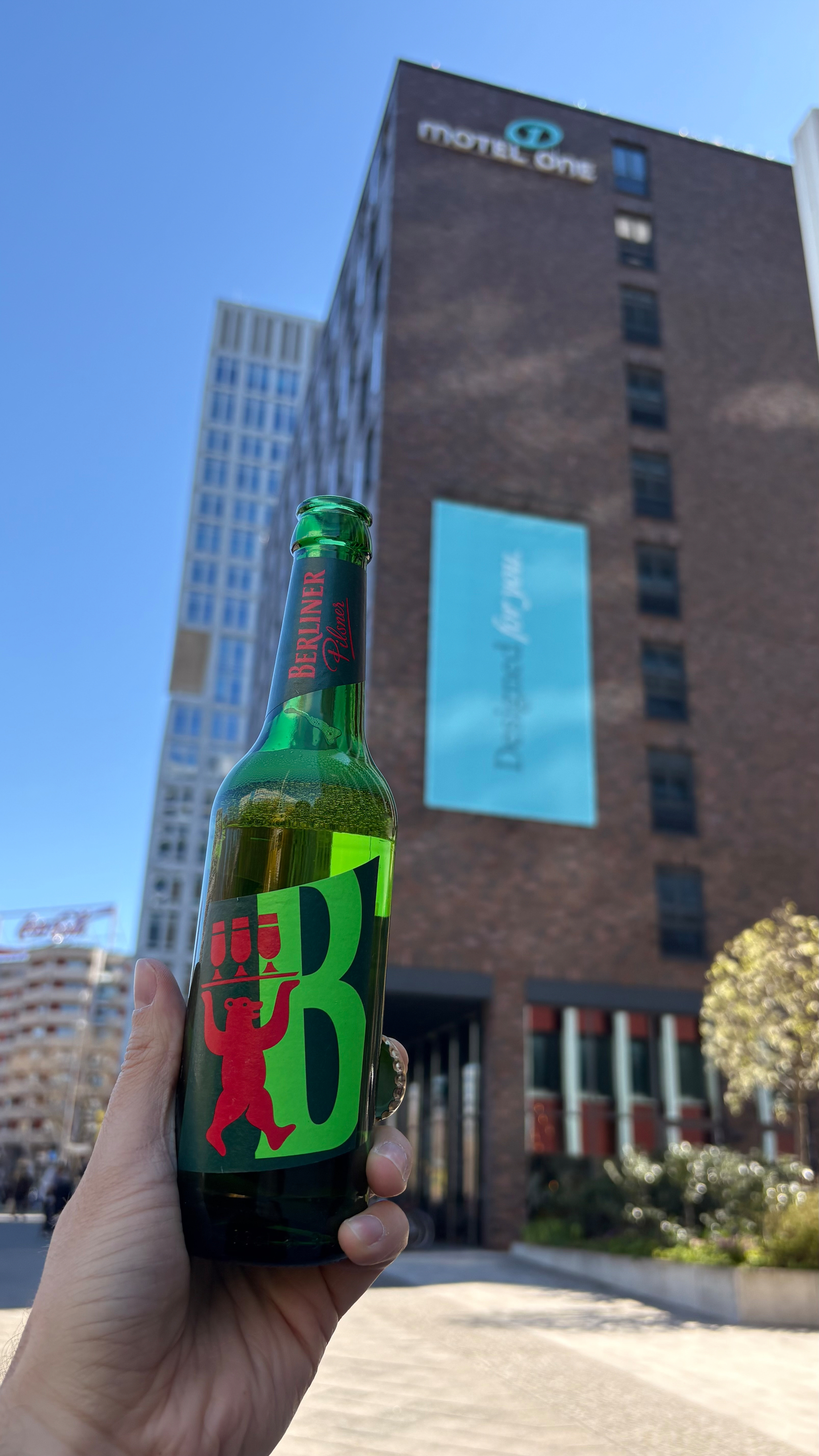 Auto-generated description: A hand holding a bottle of Berliner Pilsner is pictured in front of a modern building labeled with moxy & residence inn under a clear blue sky.