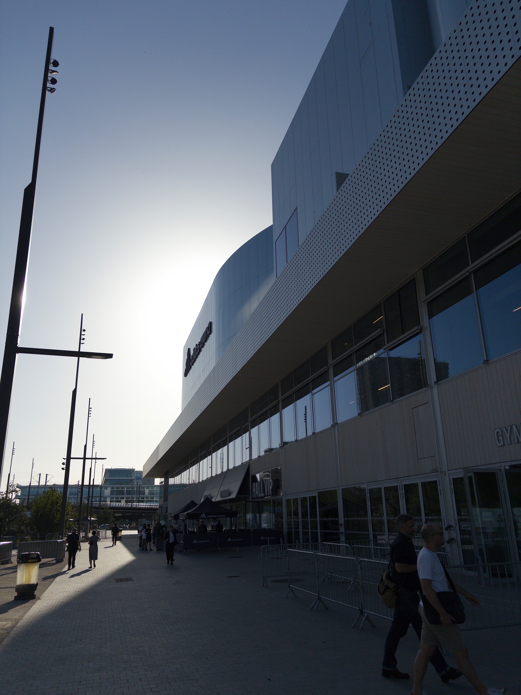 People walk along a paved path next to a modern building with reflective windows, silhouetted by the bright sun behind them.