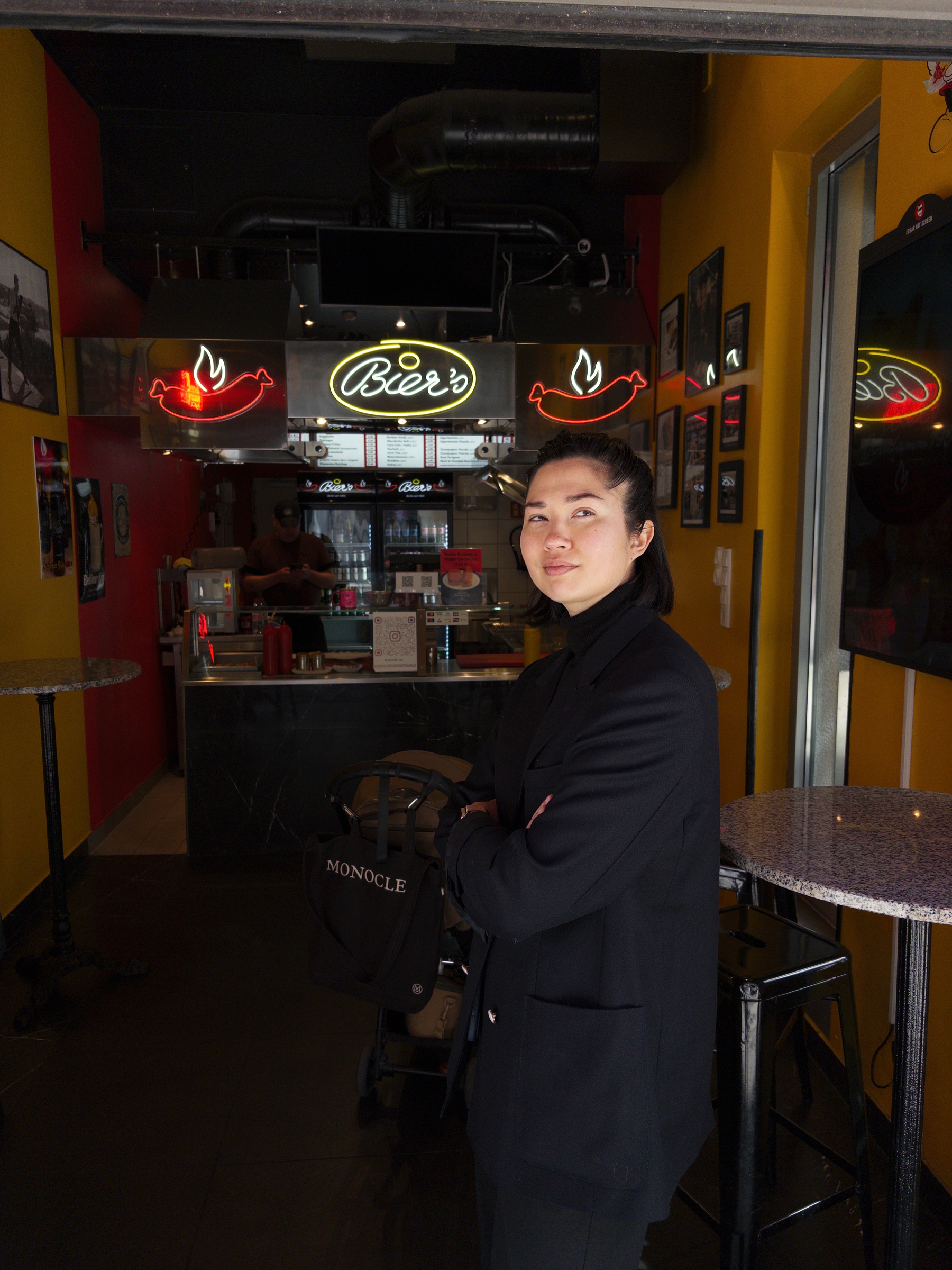 Auto-generated description: A person stands in a restaurant with colorful walls, under a neon sign that says Beer, with their arms crossed and a backpack labeled Monocle.