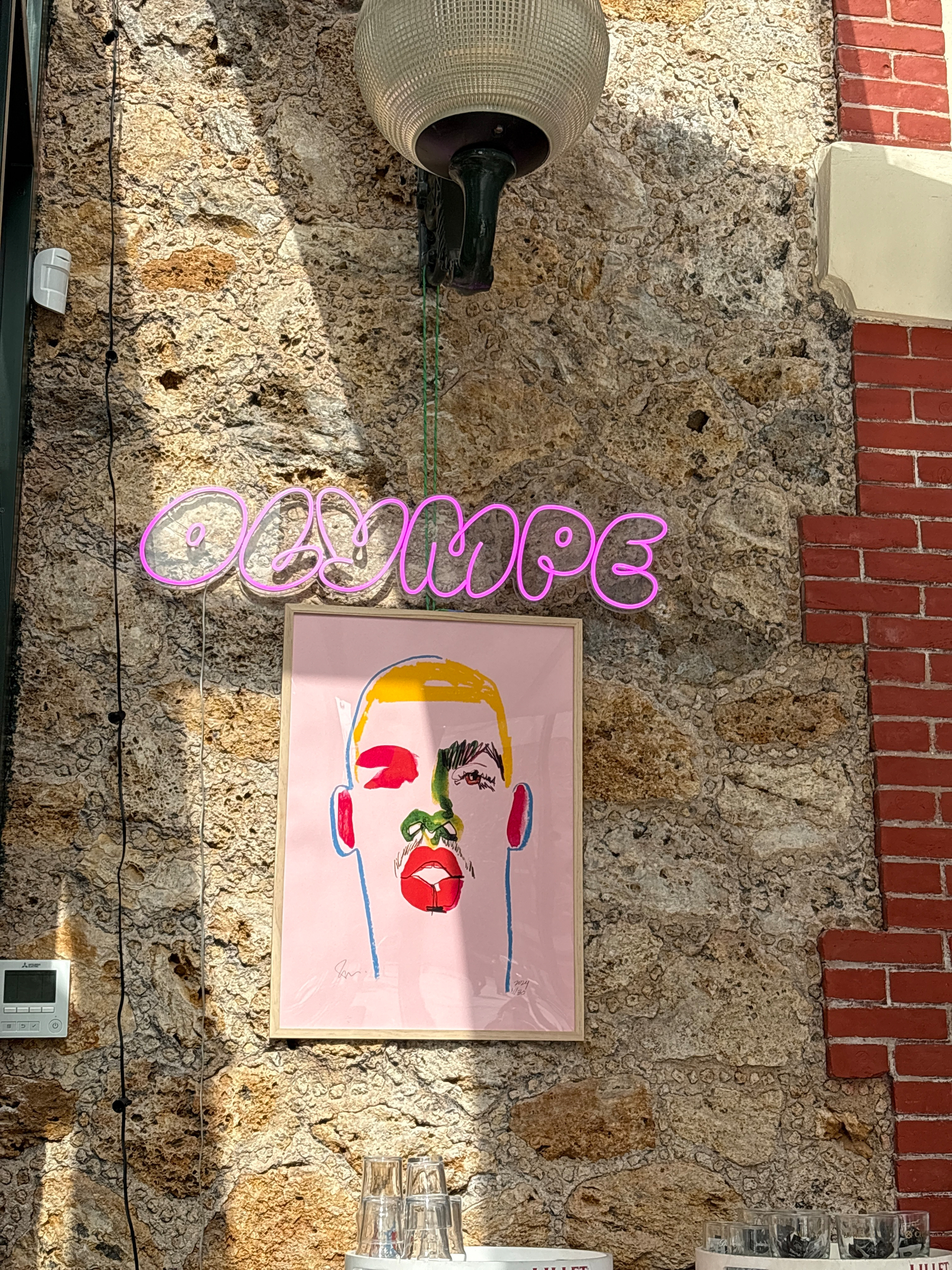 A bright, abstract portrait is framed on a textured stone wall with a neon sign above it spelling Olympe in pink letters.