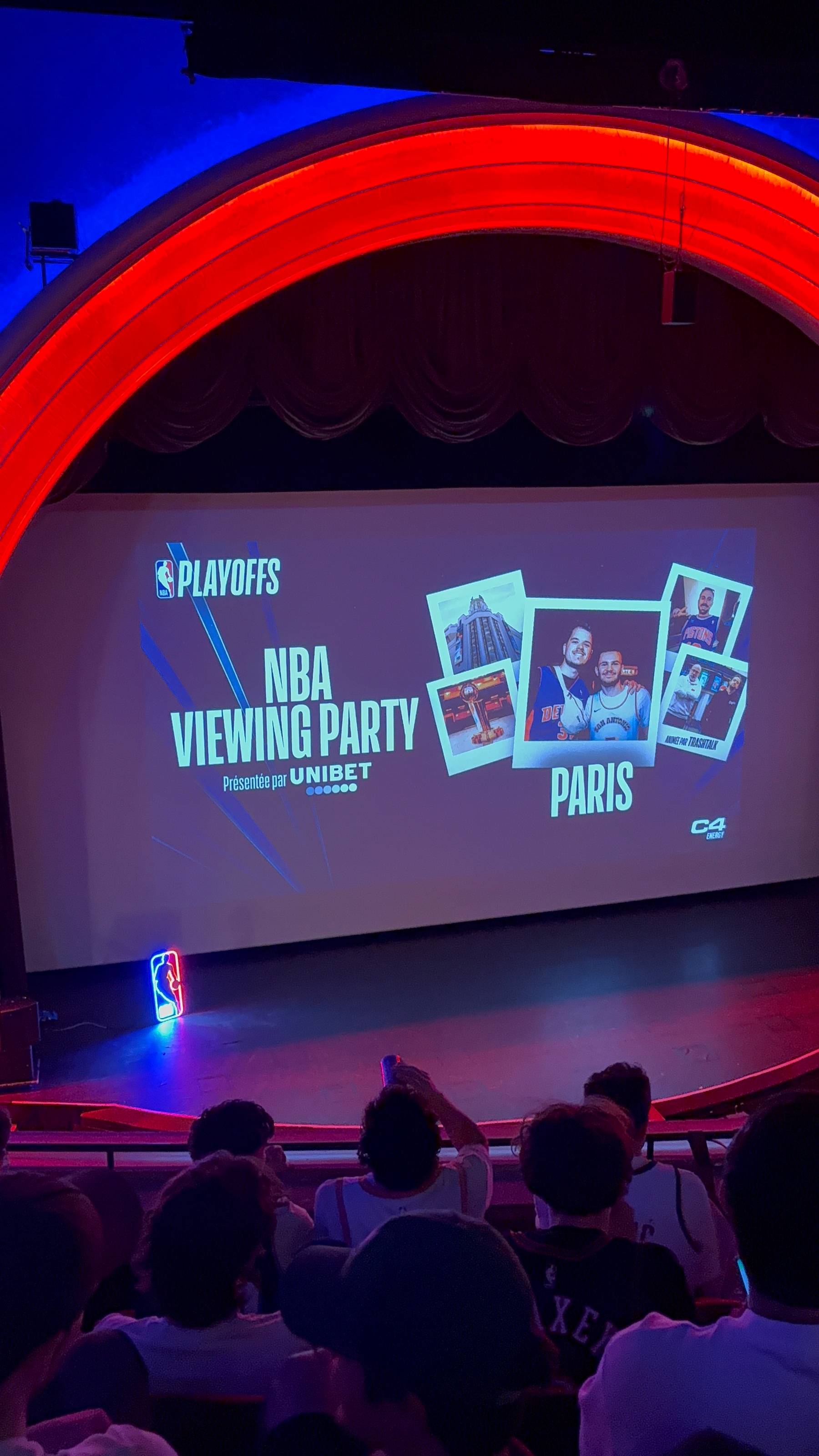 A group of people is watching a large screen promoting an NBA Playoffs viewing party in Paris presented by Unibet.