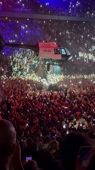 A helicopter is flying above a densely packed crowd at a concert or large event, with many people holding up their phones.