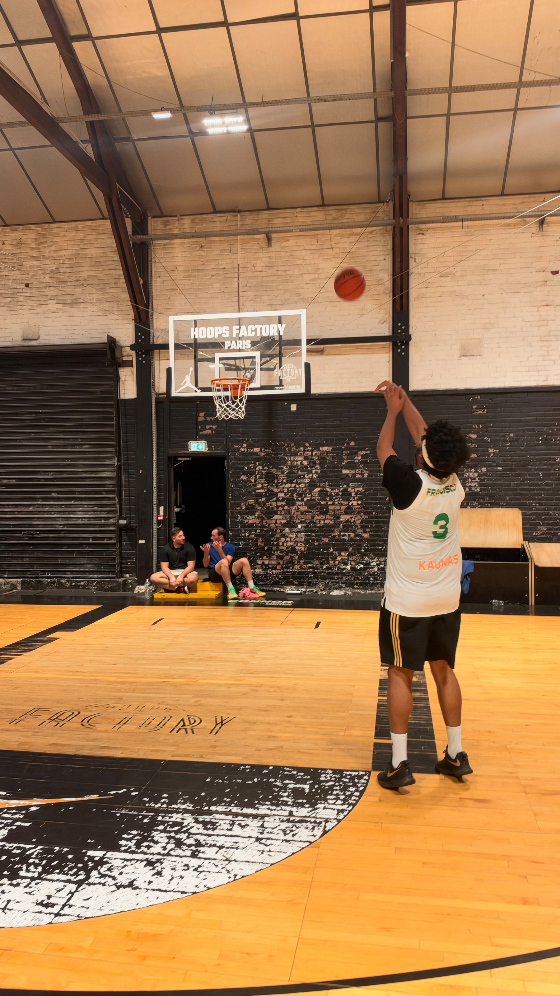 A person wearing a white basketball jersey with the number 3 is shooting a basketball towards a hoop inside an indoor court labeled HOOPS FACTORY PARIS, while two others sit against the wall in the background.