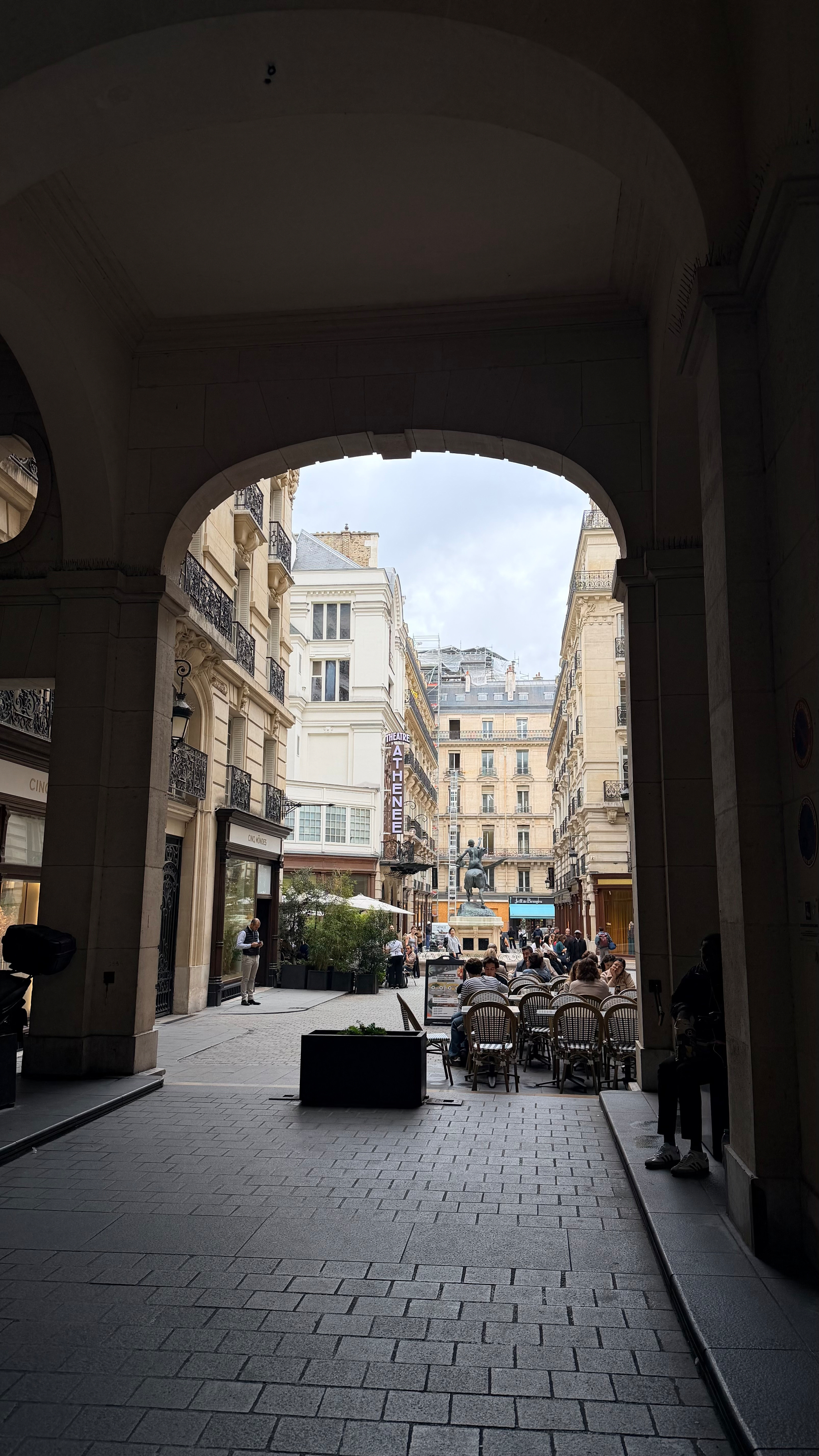 Auto-generated description: A street scene shows people seated at an outdoor café under historic buildings, viewed through a large archway.