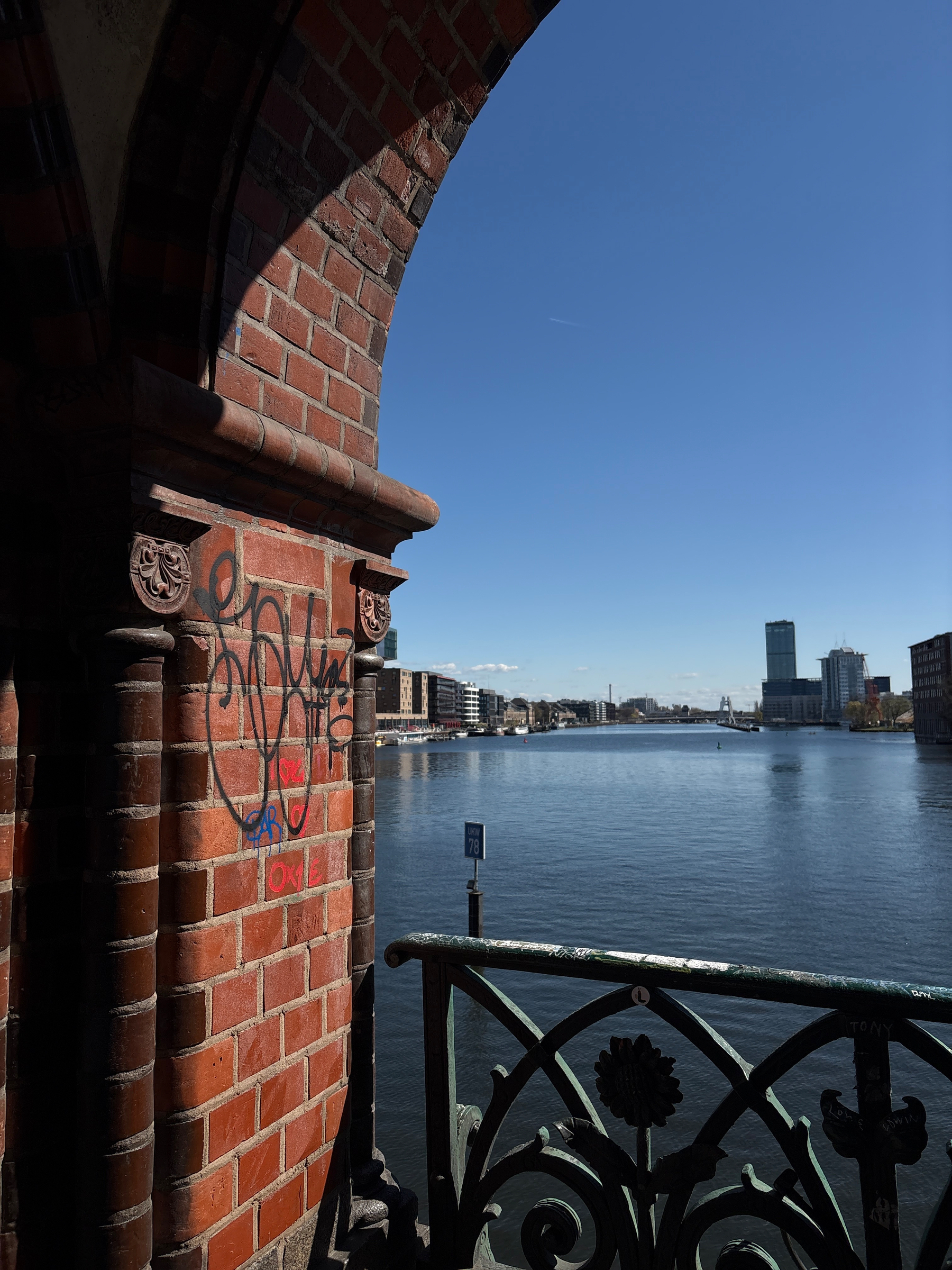 Auto-generated description: A view of a river with city buildings in the distance, framed by a brick archway and balcony with graffiti.