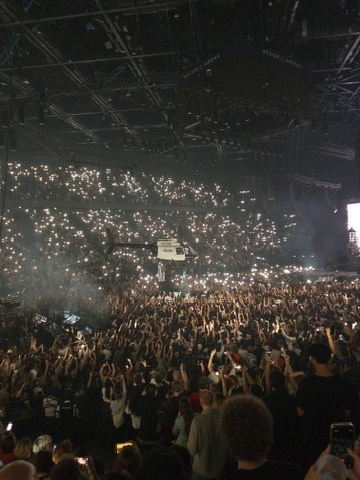 A large crowd at a concert or event is holding up their phones with the flashlights on, creating a sea of lights in a darkened arena.