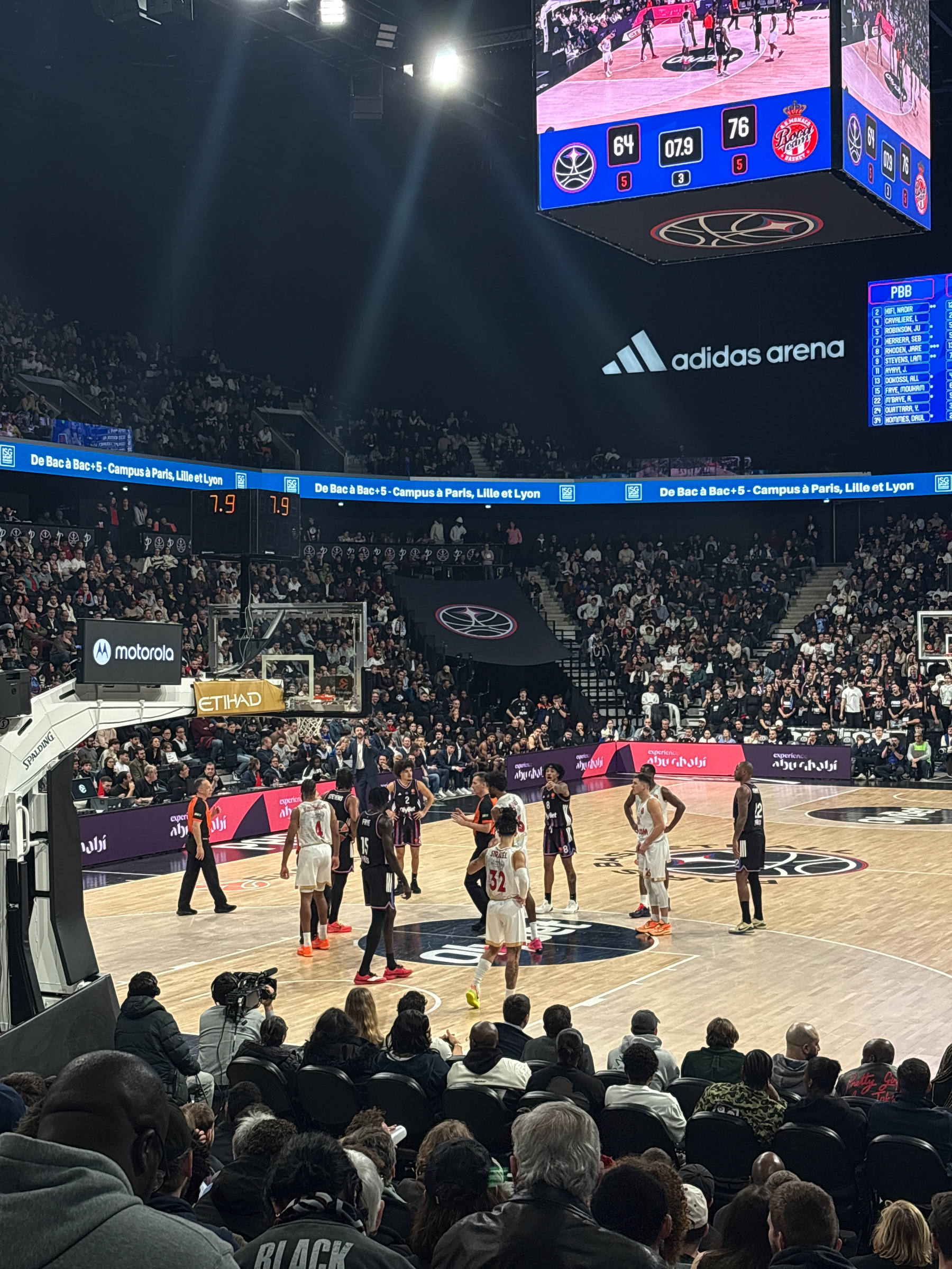 A basketball game is taking place in a crowded arena with the scoreboard displaying a score of 64 to 76.
