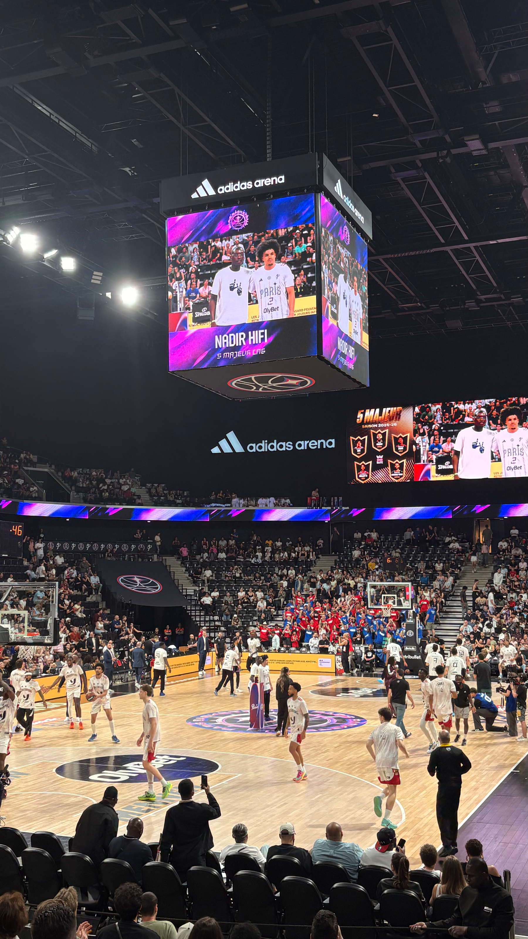 A basketball game is taking place in an indoor arena with players warming up on the court and spectators watching from the stands.