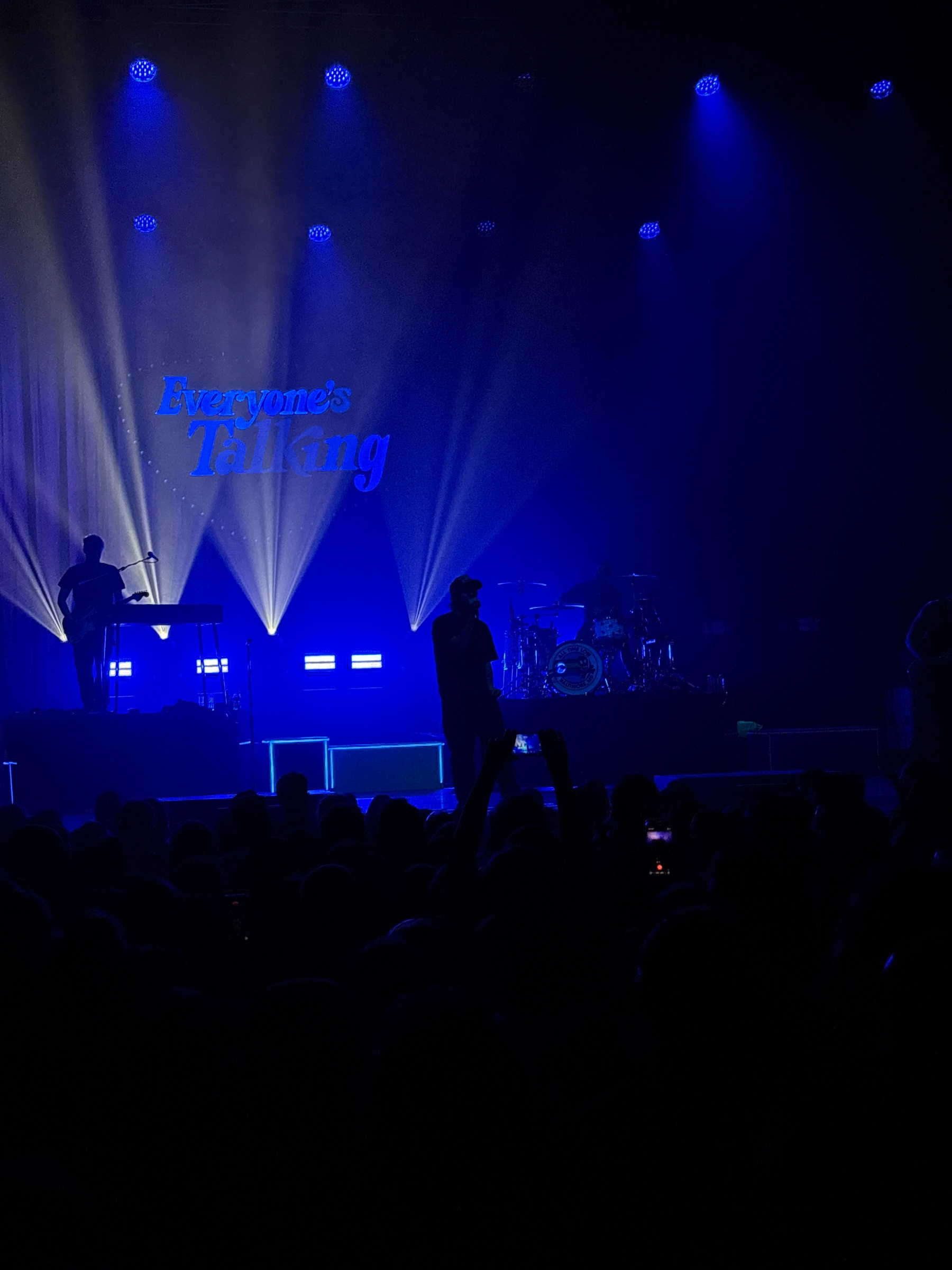 A band is performing on a dimly lit stage with blue lighting and a crowd in the foreground, featuring a backdrop that says Everyone’s Talking.