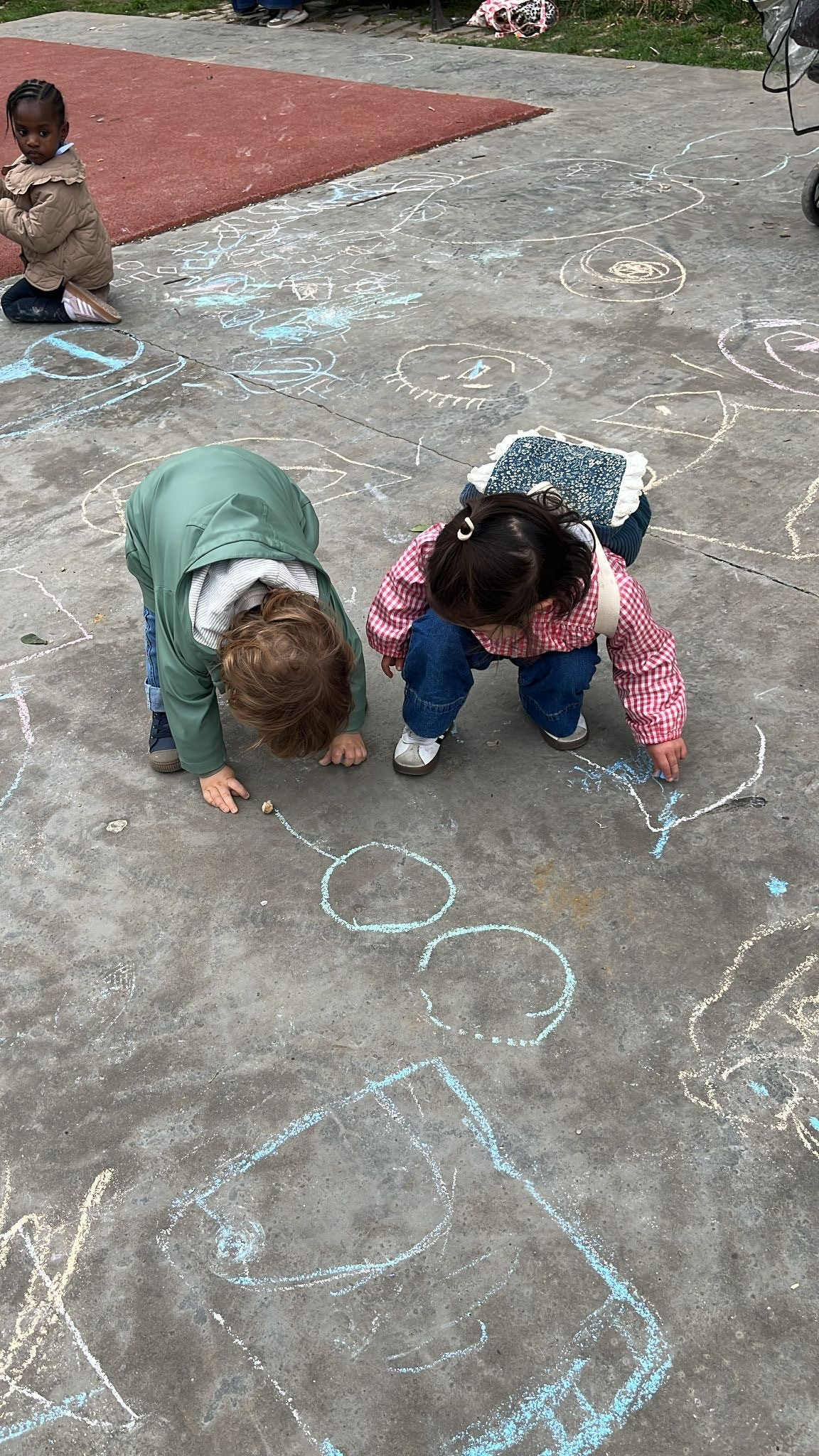 Two children are drawing with chalk on the pavement while other kids and drawings are in the background.