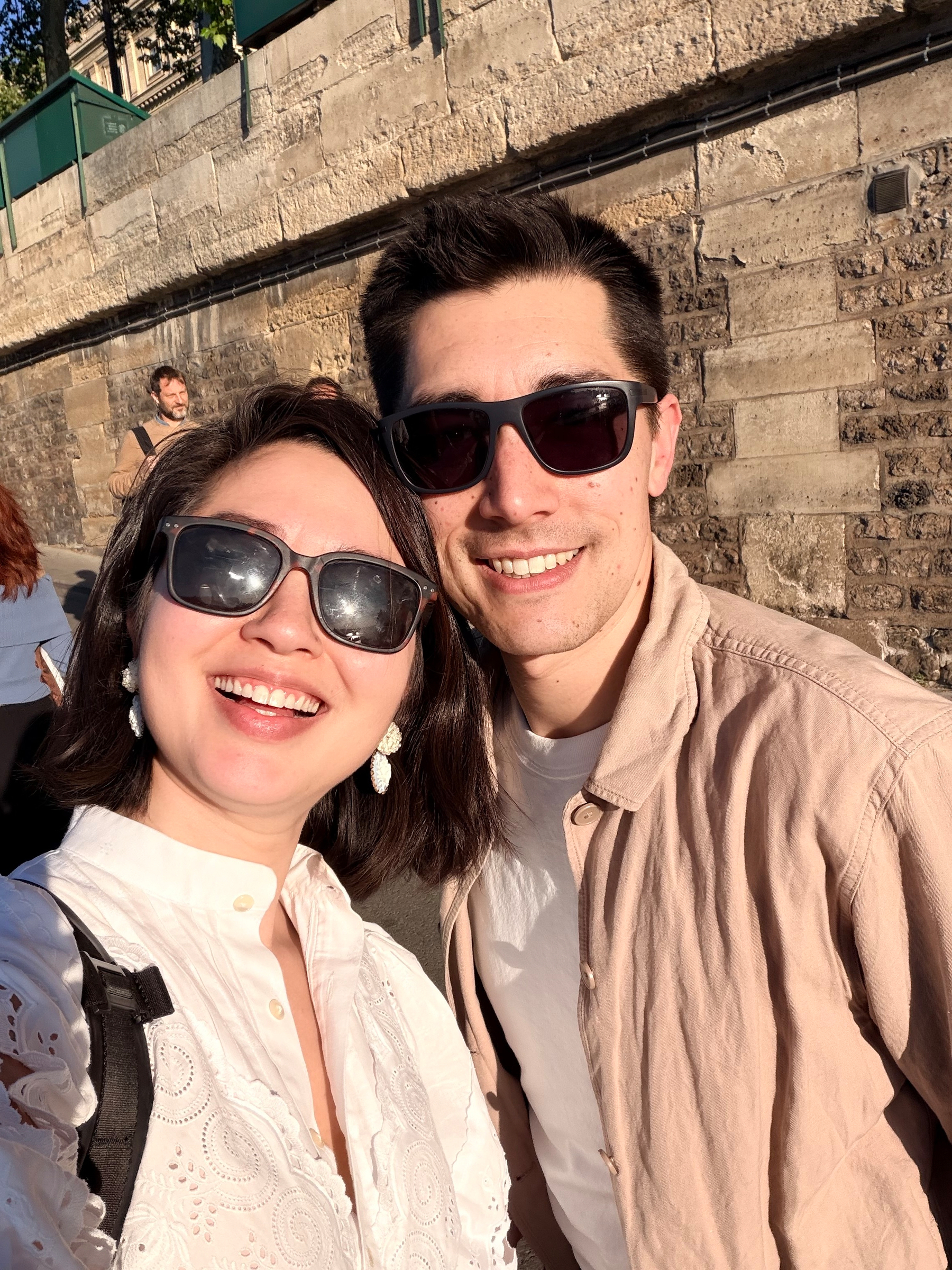 A smiling couple wearing sunglasses enjoys a sunny day in front of a stone wall.