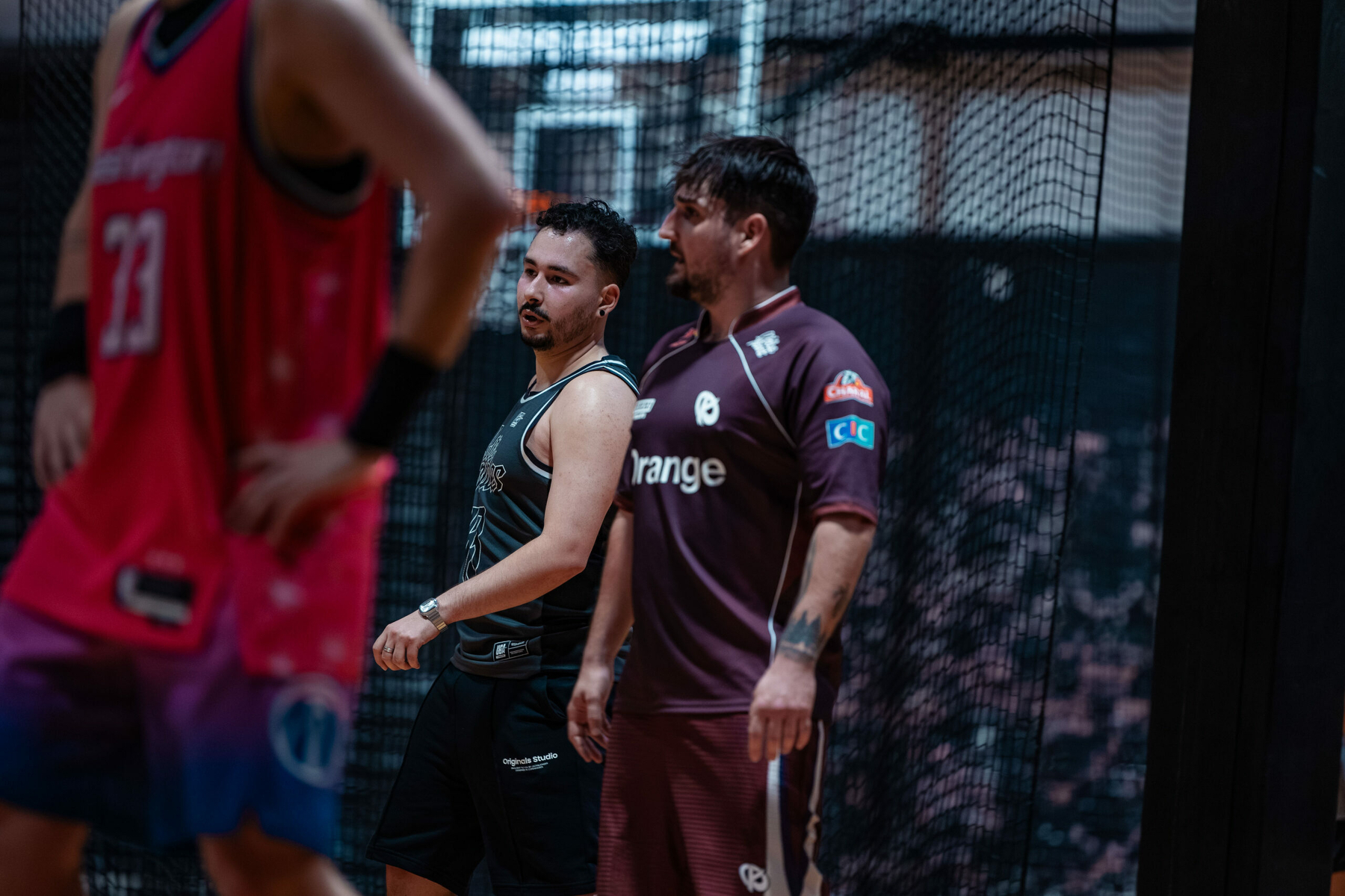Auto-generated description: Two men in sports attire are standing on a basketball court, with one wearing a black jersey and the other in a brown jersey, as another person in a pink jersey is partially visible.