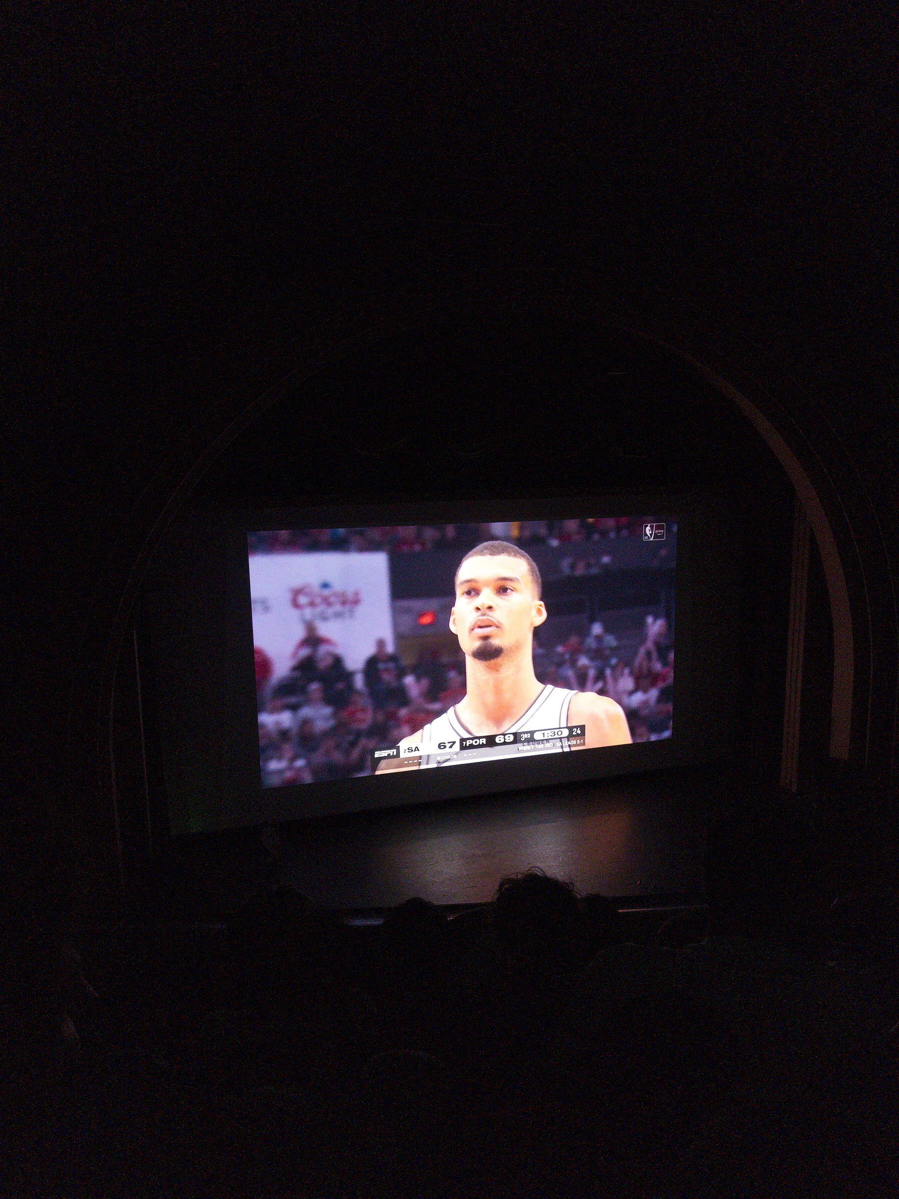 A large screen in a dimly lit theater displays a close-up of a basketball player during a game.