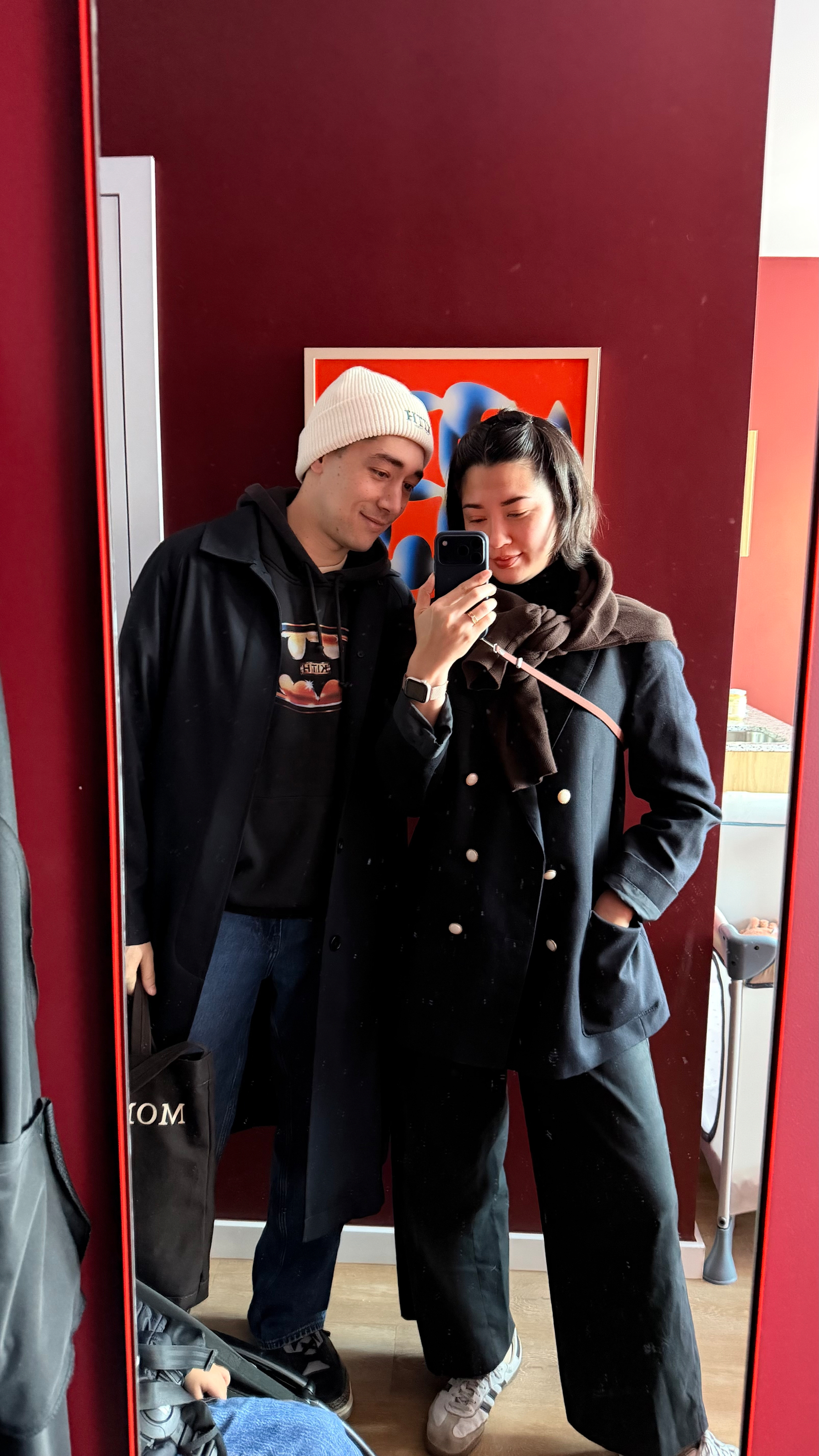 Auto-generated description: A casually dressed couple is posing for a mirror selfie in a room with red walls.