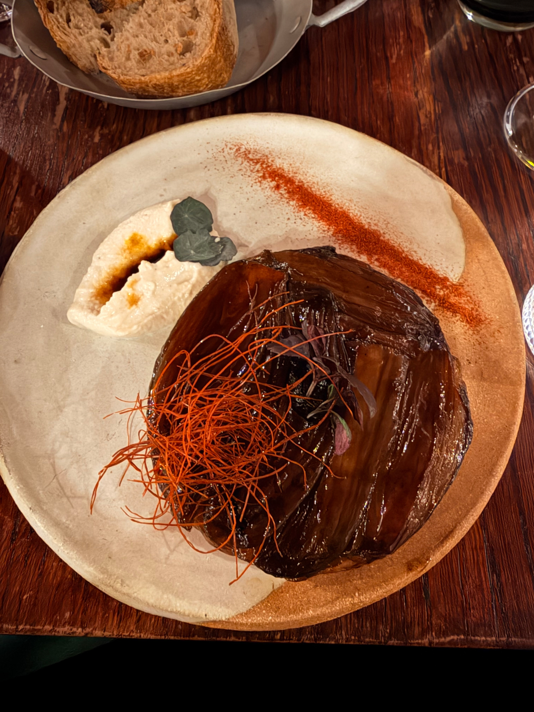 A gourmet dish featuring a glazed vegetable topped with fine red strands is artfully presented alongside creamy sauce and bread on a textured plate.