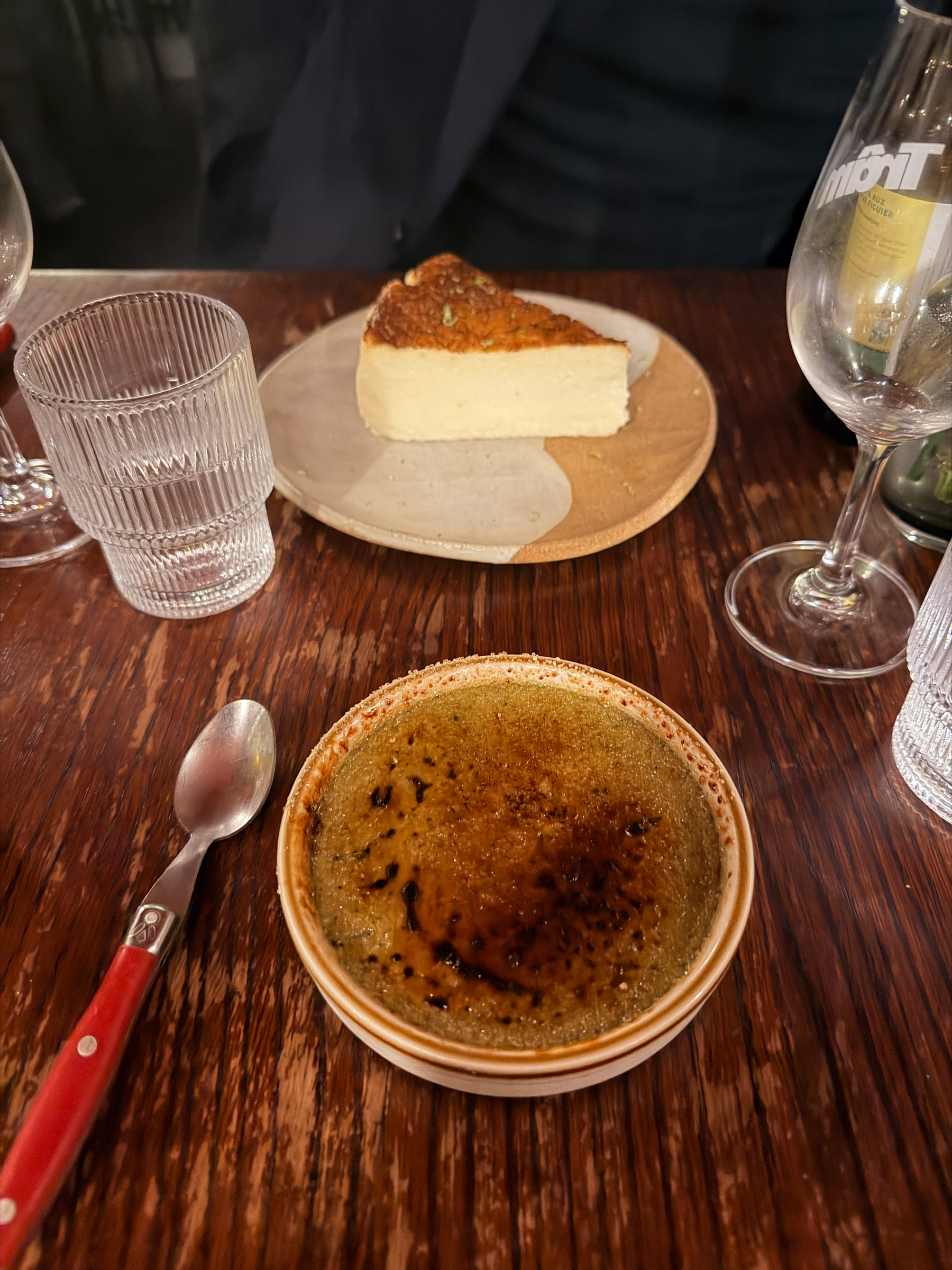 A table features a plate with cake, a glass of wine, a glass of water, and a dish, possibly crème brûlée, with a red-handled spoon beside it.