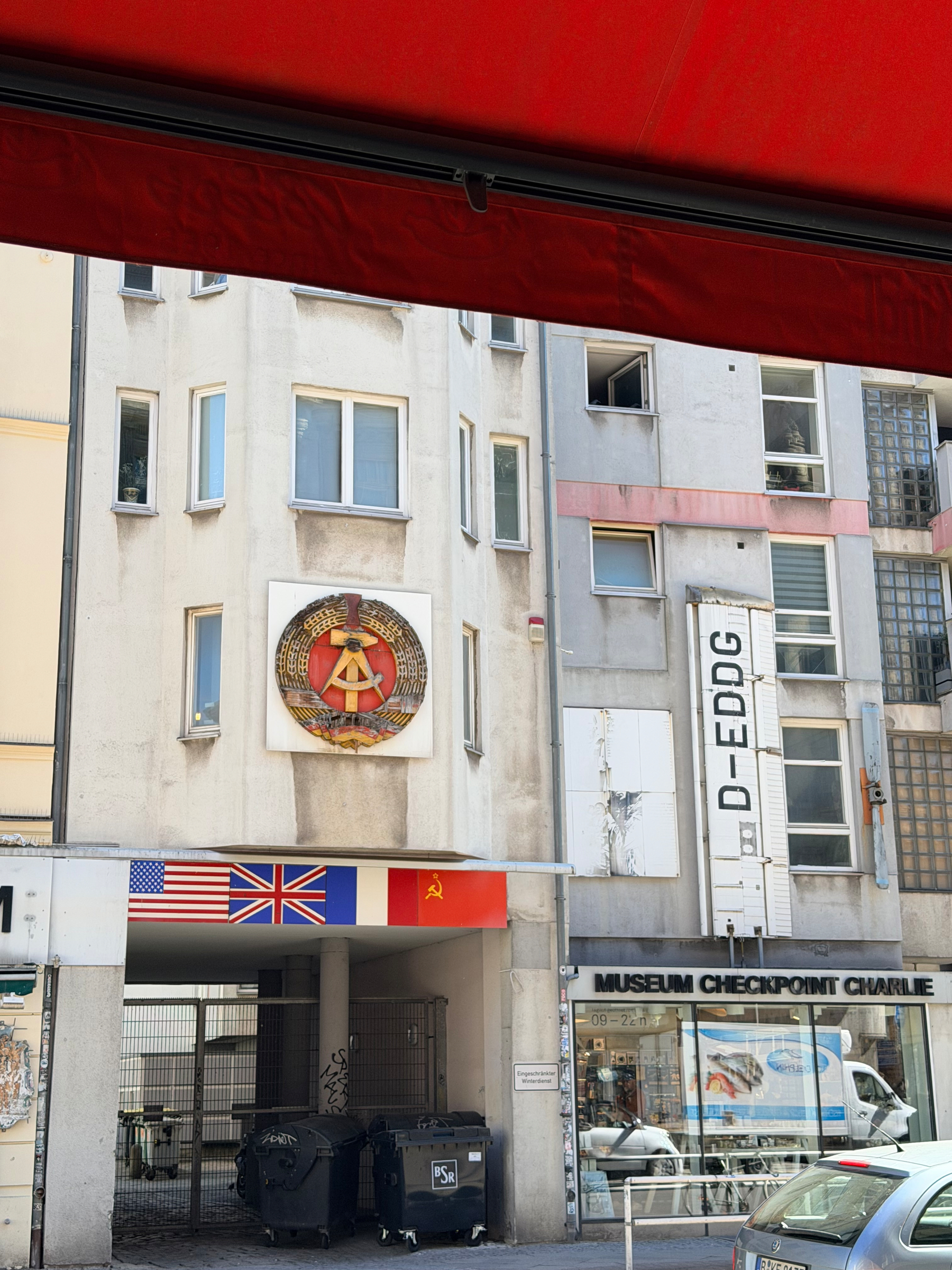 Auto-generated description: A building with a prominent emblem on its facade stands above a tourist site featuring flags of the United States, United Kingdom, and the Soviet Union near Museum Checkpoint Charlie.