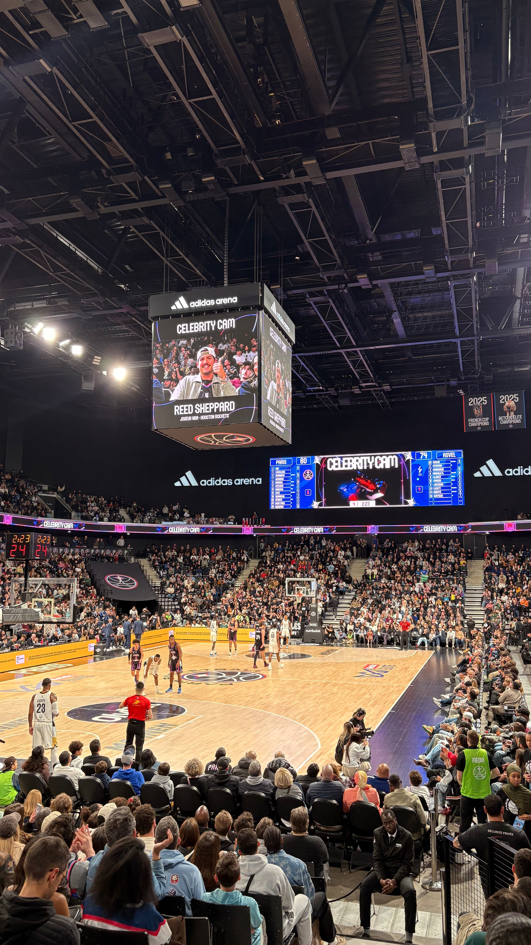 A crowded basketball arena features a live game with people seated around the court and large screens displaying player information.