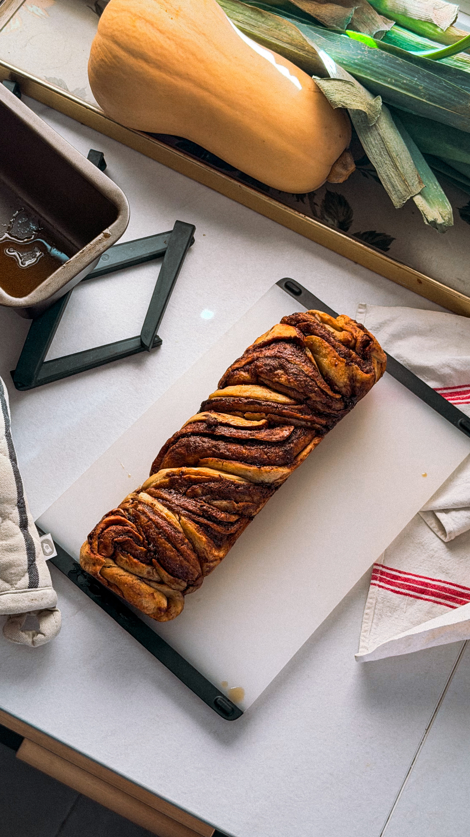 Auto-generated description: A freshly baked twisted cinnamon bread loaf is placed on a white cutting board next to a butternut squash, a baking pan with some leftover syrup, and kitchen towels on a countertop.
