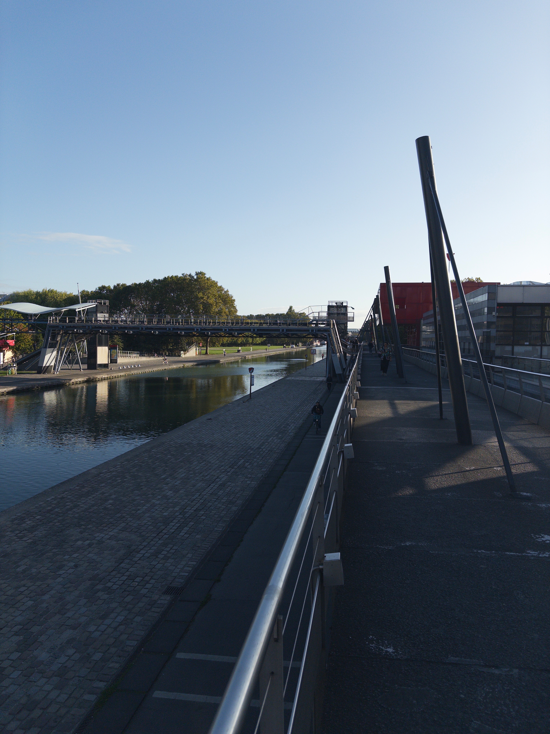 A pedestrian bridge runs alongside a calm river with trees and buildings visible in the background under a clear sky.