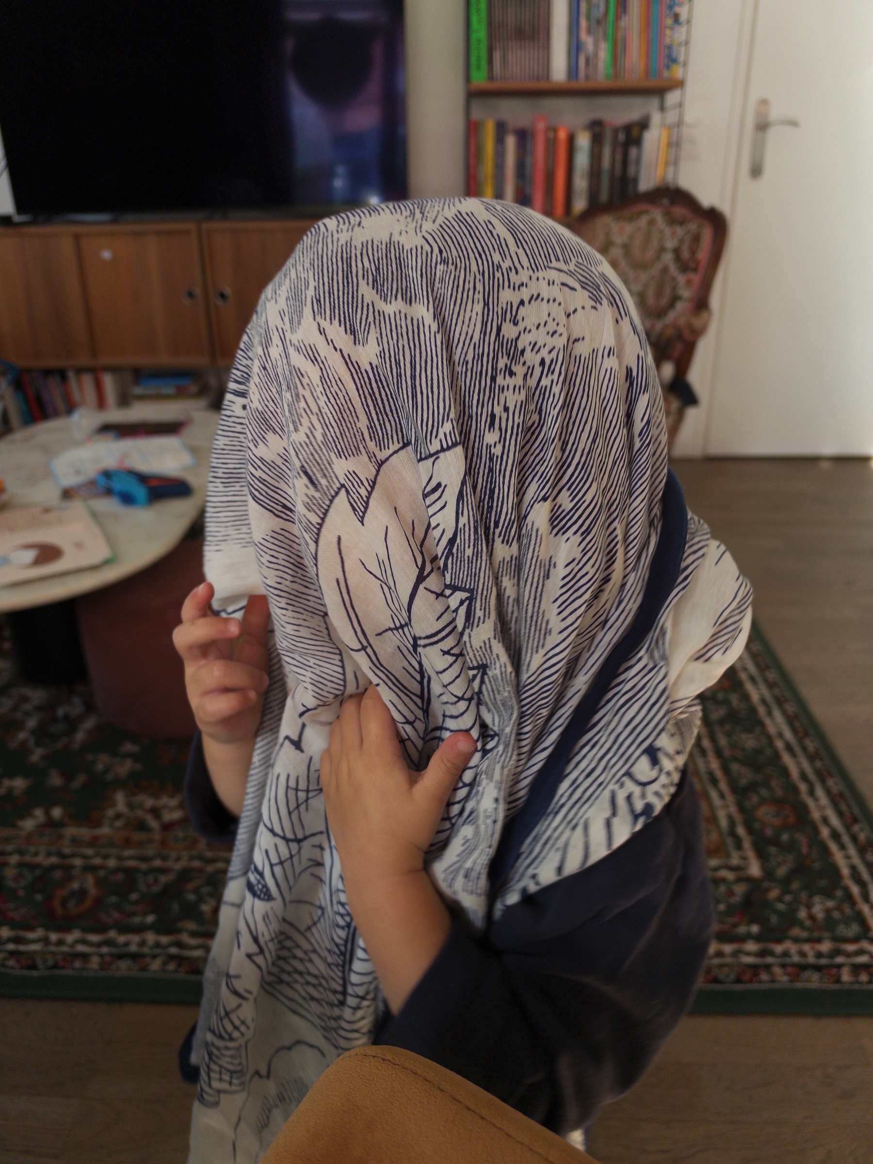 Auto-generated description: A child is playfully covering their head with a patterned cloth inside a living room.