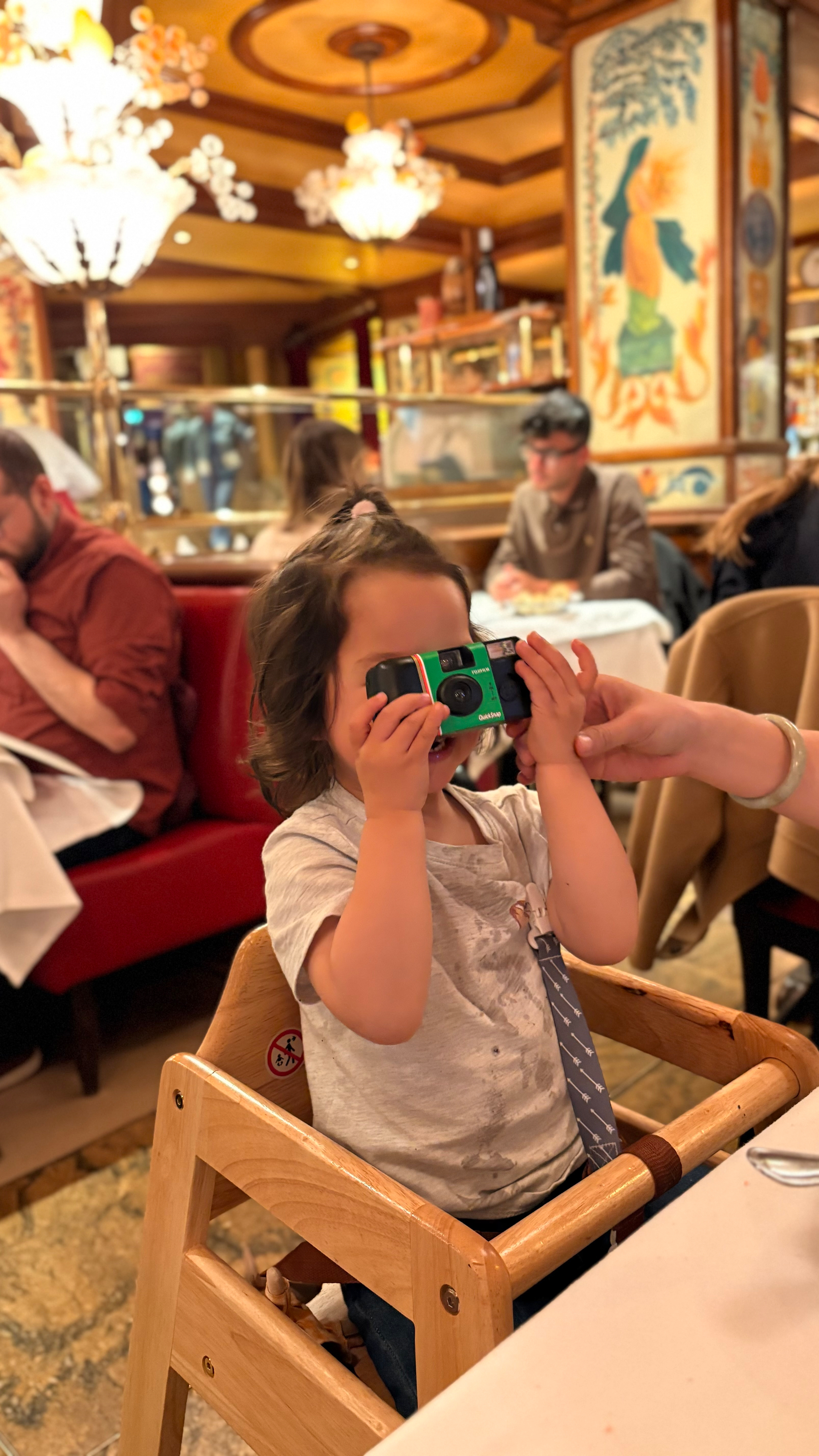 A young child sitting in a high chair is holding a camera to their face in a restaurant with a warm and colorful interior.