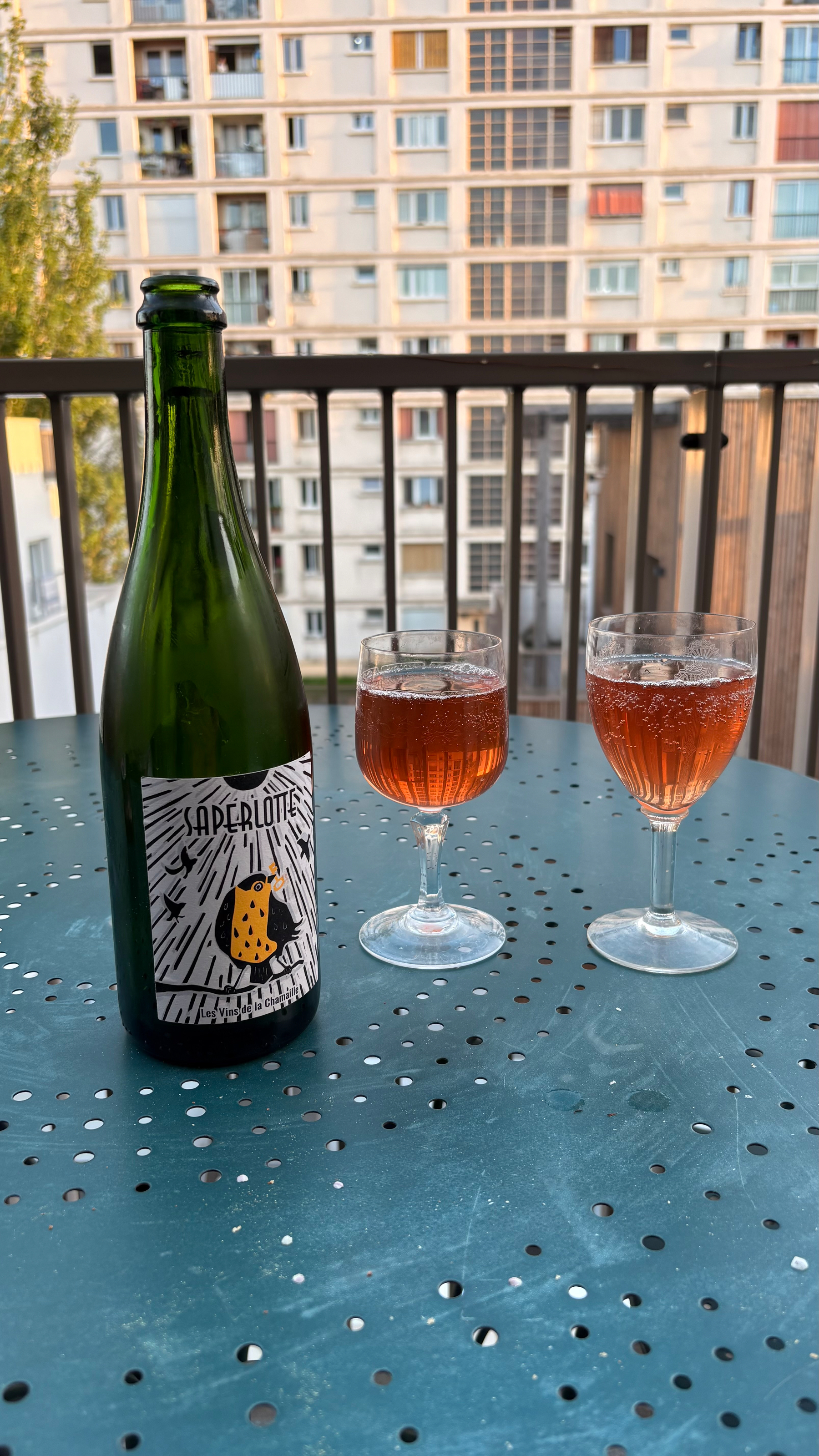 A bottle and two glasses of a pinkish drink are set on a teal perforated table on a balcony with a view of urban buildings.