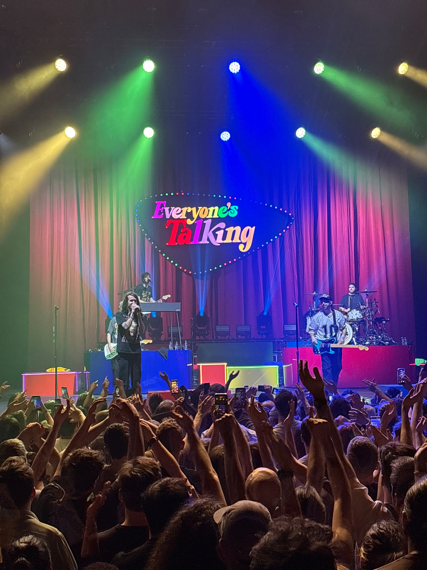 A live band performs on stage under colorful lights with a crowd cheering below and a backdrop displaying Everyone's Talking.