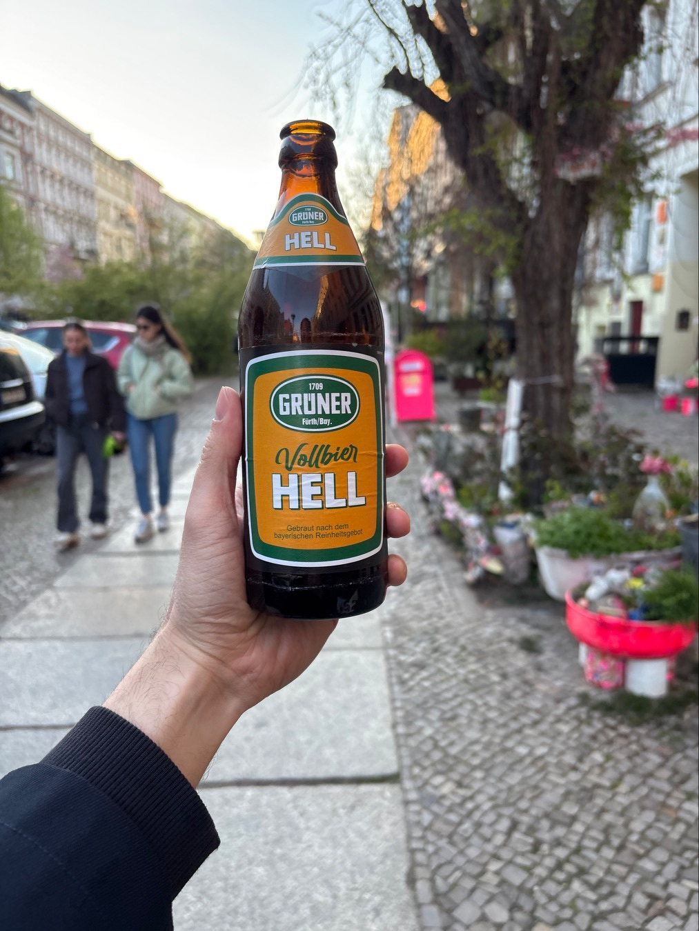 Auto-generated description: A hand is holding a Grüner Vollbier Hell beer bottle on a street lined with plants and people walking in the background.