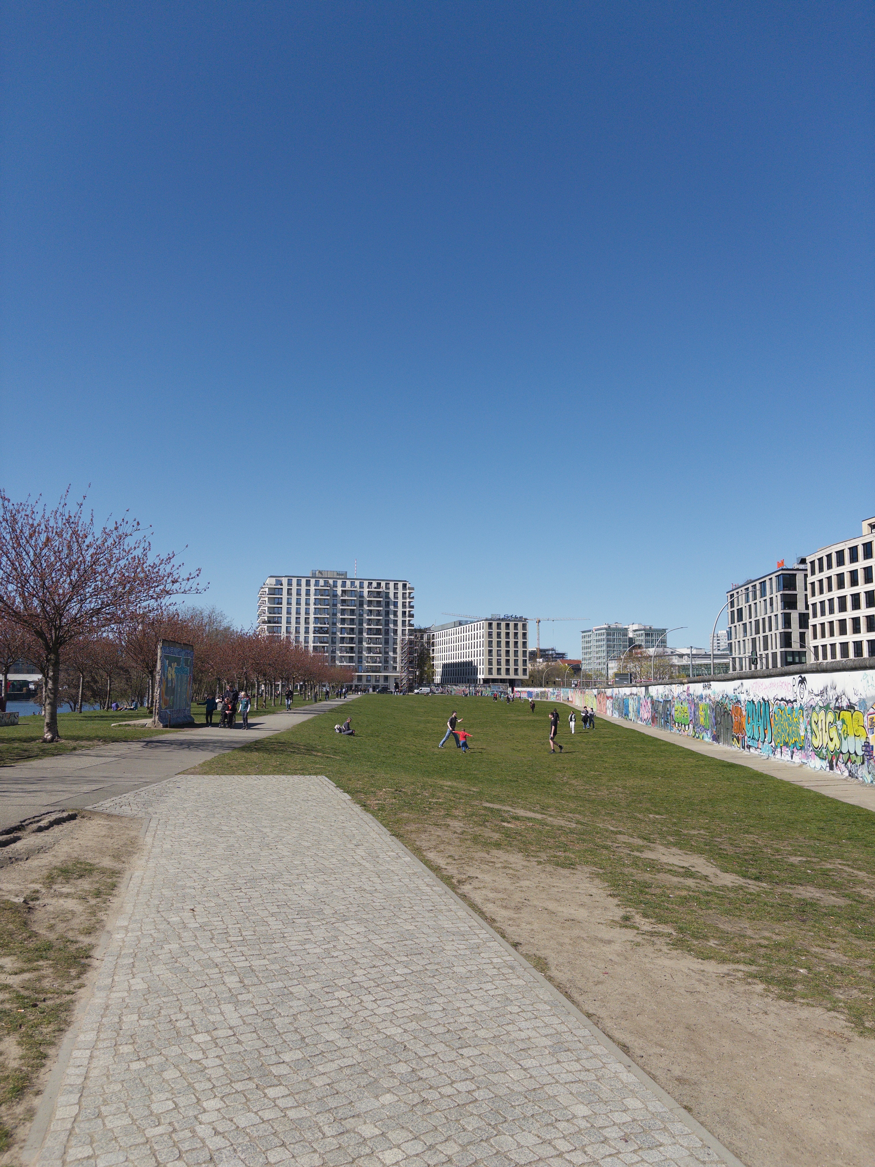 Auto-generated description: A paved path runs through a park with people relaxing on the grass, surrounded by trees, modern buildings, and graffiti-adorned walls under a clear blue sky.