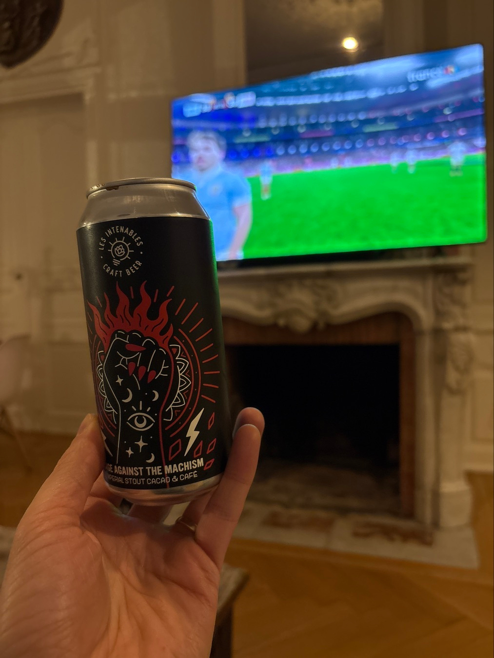 A hand holding a can of craft beer with an artistic label is in the foreground, while the background shows a soccer match on a television above a fireplace.