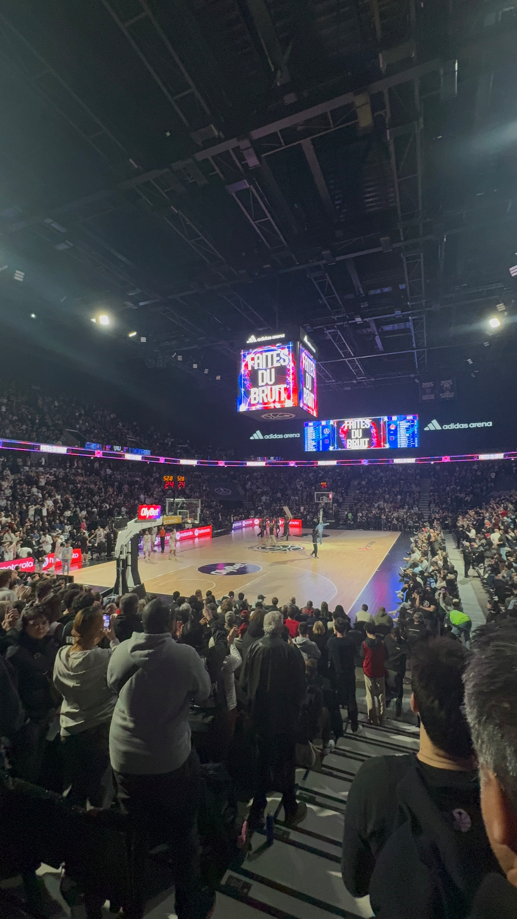 A lively basketball game is taking place in a large indoor arena filled with cheering spectators.