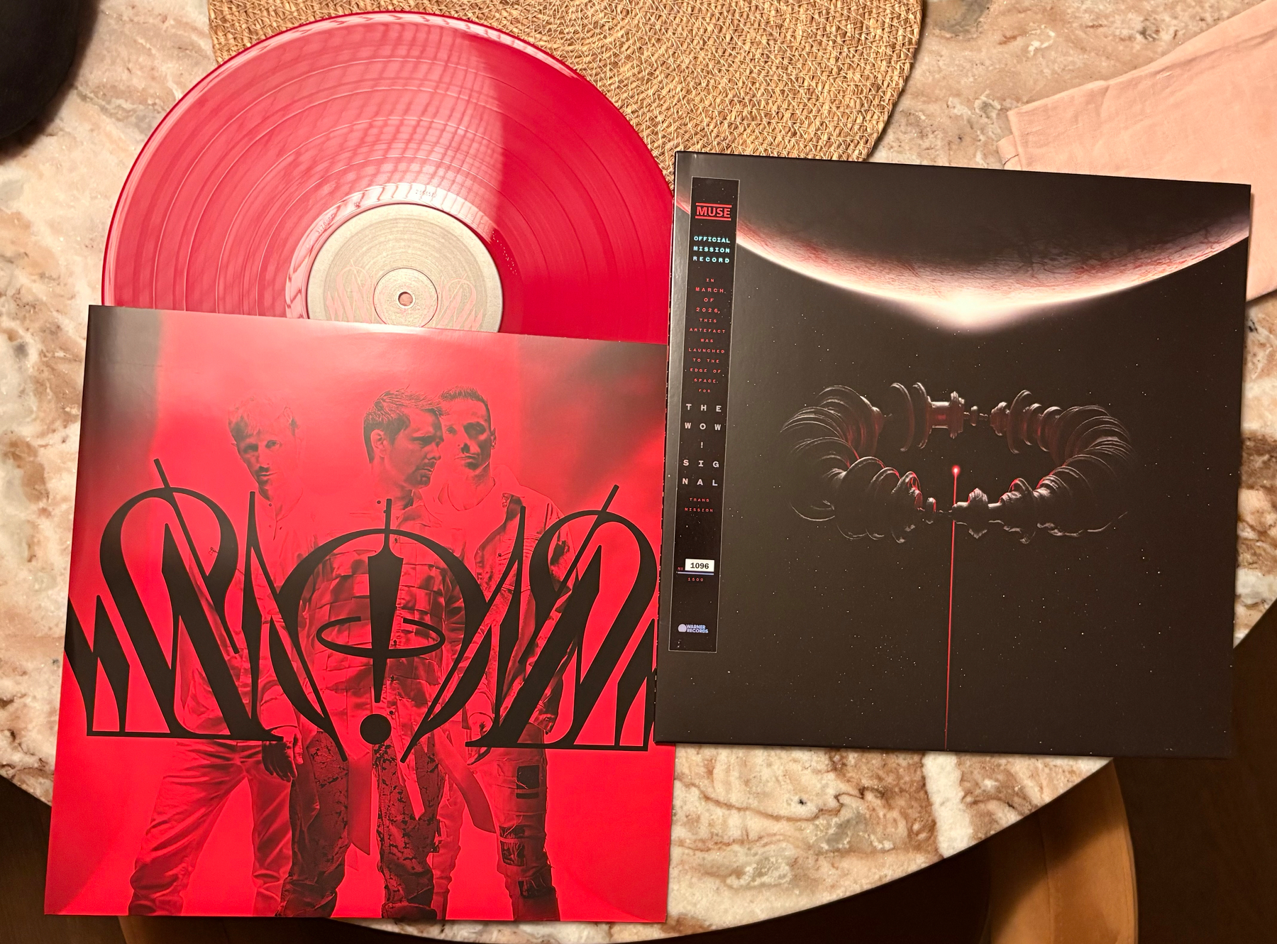 A red translucent vinyl record partially pulled from a cover featuring two men and abstract symbols lies next to a black album cover with an otherworldly design on a round table.
