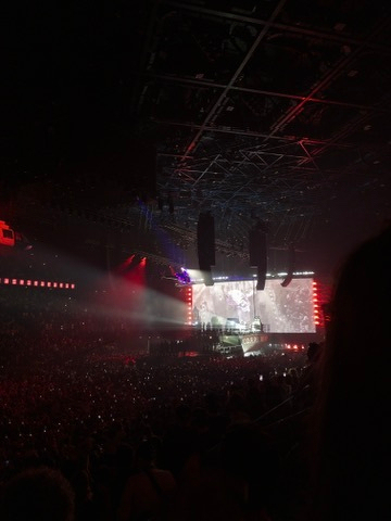 A large crowd is gathered in a dark indoor arena watching a brightly lit stage with screens and red lighting effects.